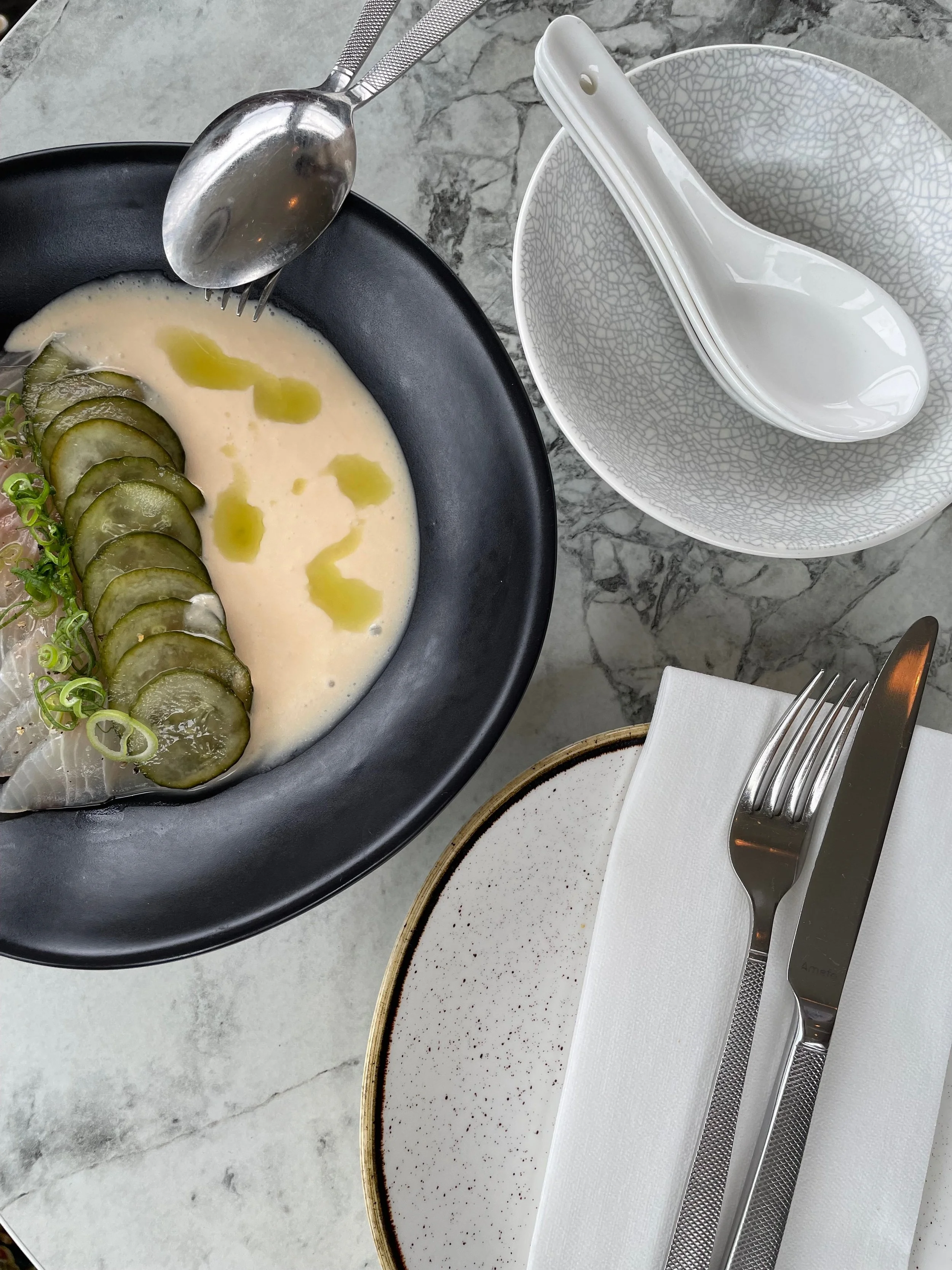 A bowl with sliced cucumbers, microgreens, and a white creamy sauce garnished with olive oil droplets, with a fork and spoon on a marble table. There are also empty white dishware including a bowl with a spoon, a plate with a napkin, fork and knife.