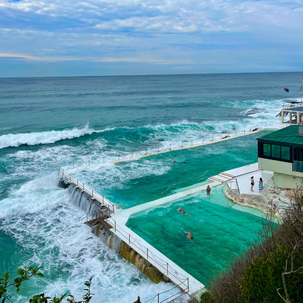 A seaside infinity pool with several people swimming and relaxing, located next to the ocean with visible waves and a cloudy sky above.