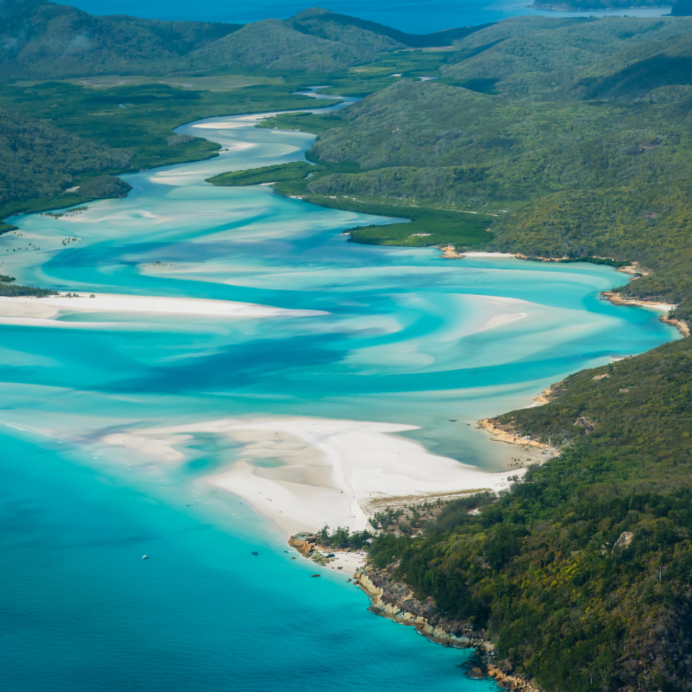 Aerial view of a winding river with blue and white sandbars, bordered by green mountains and coastline.