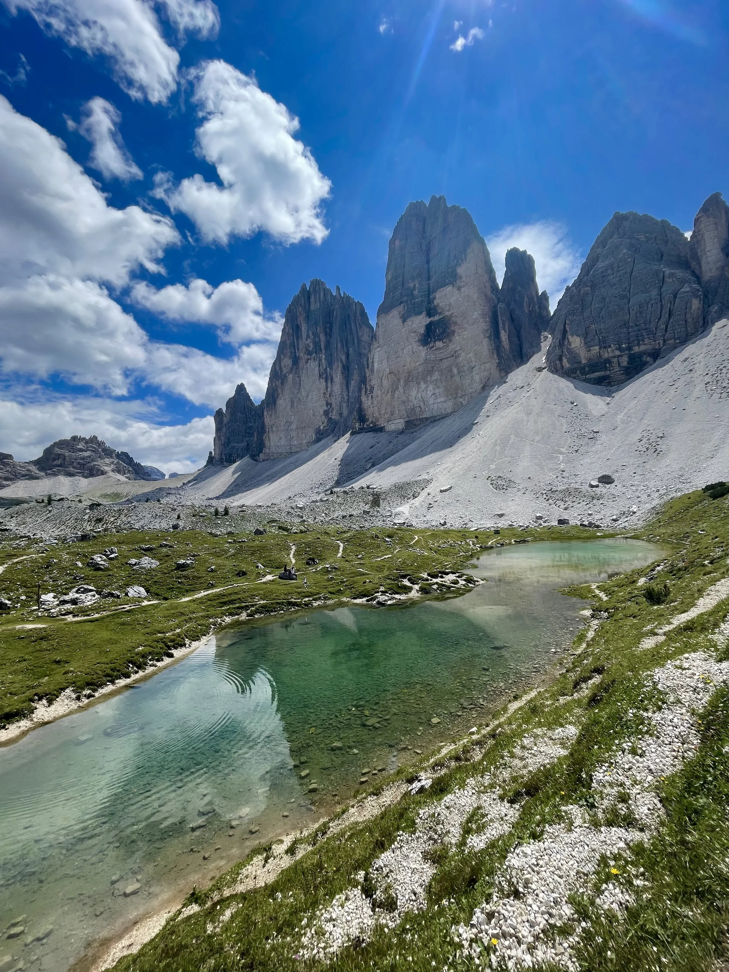 A scenic mountain landscape with towering rocky peaks, a small greenish lake reflecting the sky, and a partly cloudy sky overhead.