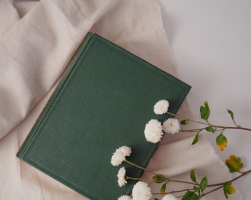 A green hardcover book resting on a cream-colored fabric with white flowers and green leaves placed on top.