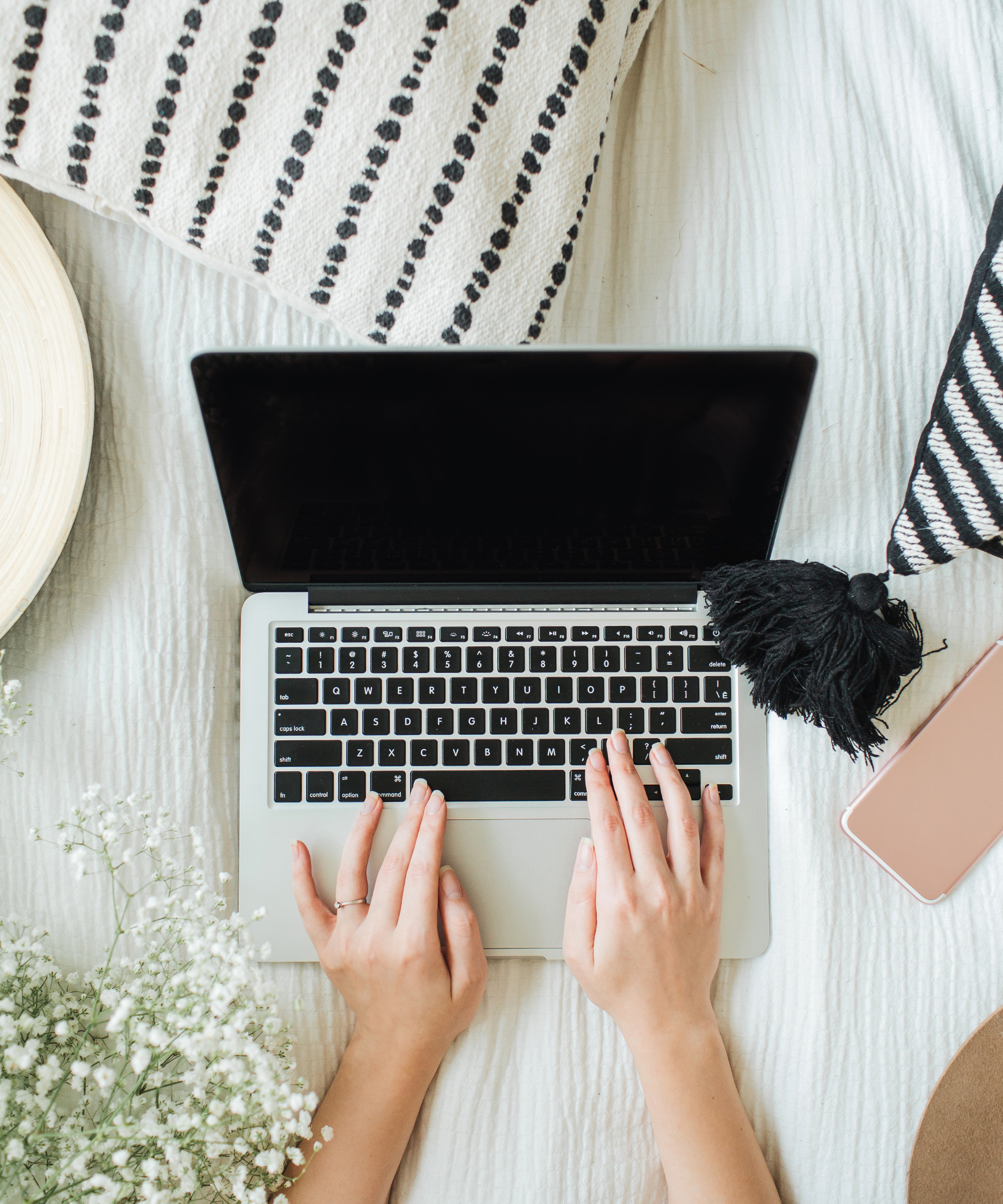 Top view of a person typing on a silver laptop with a black screen, surrounded by a pink phone, a black and white hat, a black and white striped towel, a beige plate, and white flowers on a textured cream-colored surface.