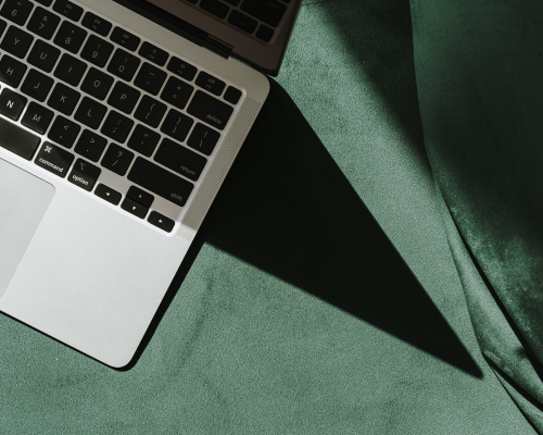 Top-left corner of a silver laptop with black keys on a green fabric surface, casting a shadow.