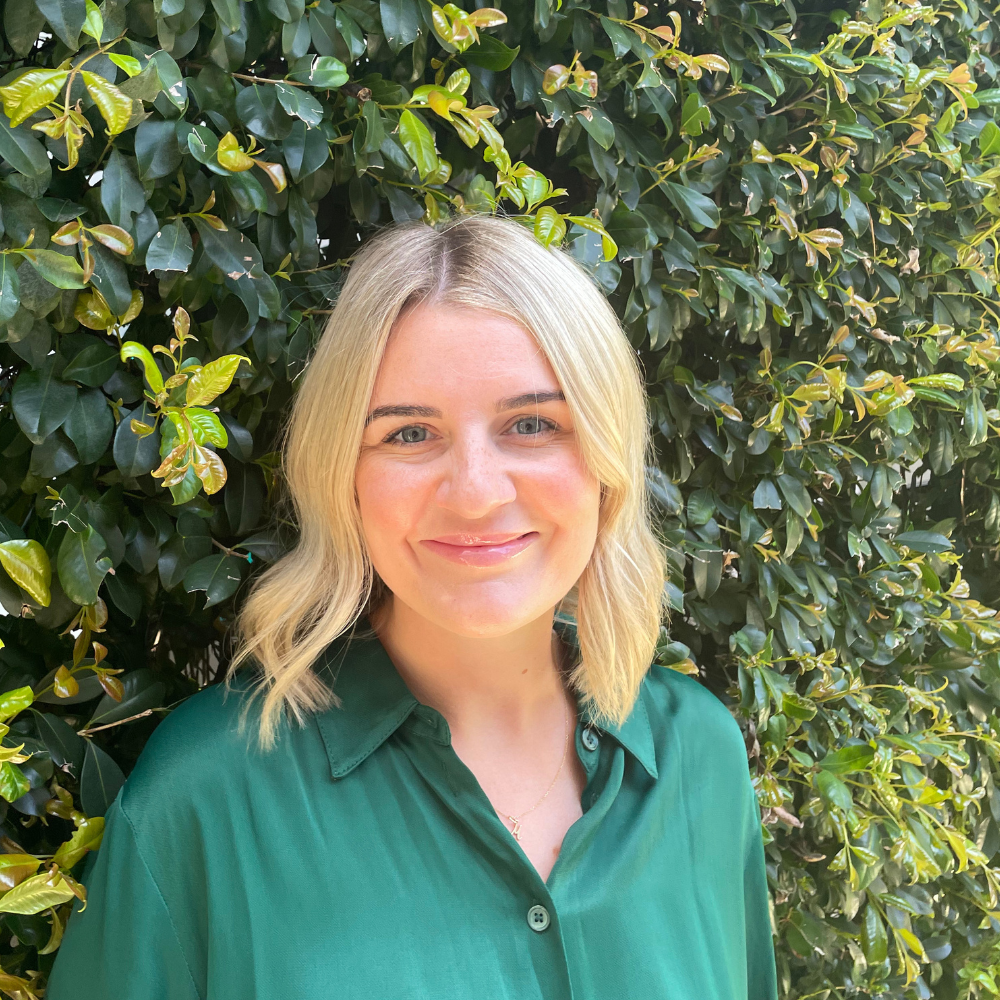A woman with shoulder-length blonde hair smiling, wearing a dark green button-up shirt, standing in front of a lush green leafy bush.