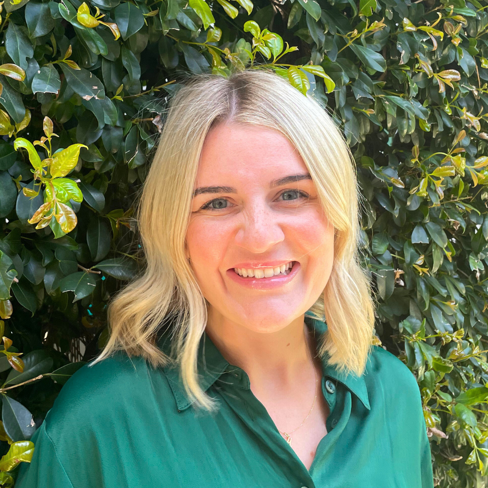 A smiling woman with blonde hair wearing a green shirt standing in front of green leafy bushes.