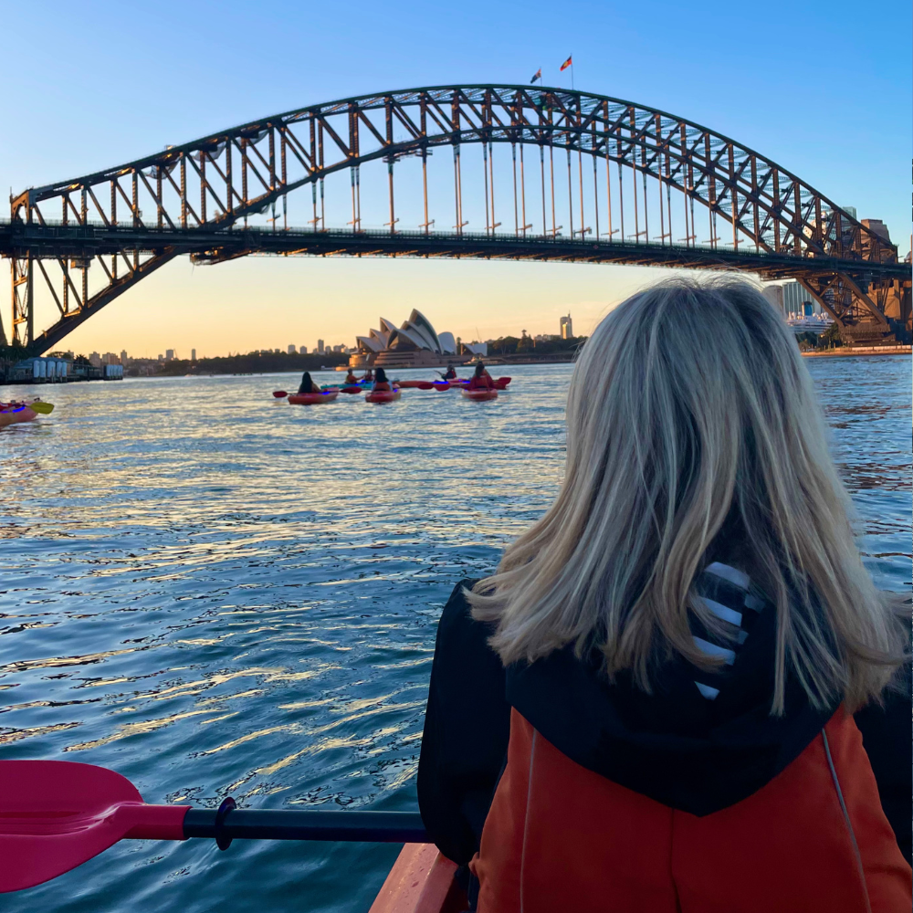 A woman with blonde hair sitting on a boat near the Sydney Harbour Bridge, with kayakers in the water and the Sydney Opera House in the background during sunset.