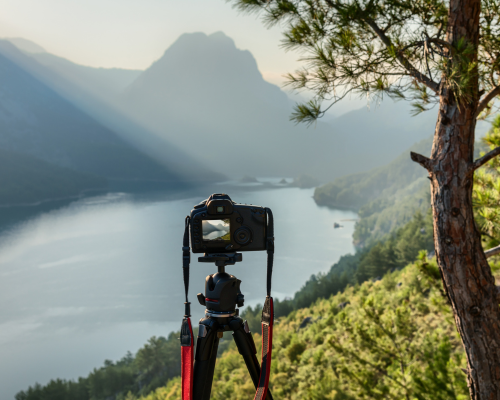 Camera on a tripod set up on a hill overlooking a river and mountains with a tree to the right, captured during daylight hours.