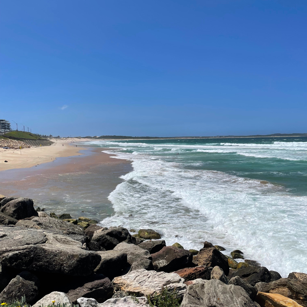 View of a sandy beach with rocks in the foreground, waves crashing on the shore, and a clear blue sky.