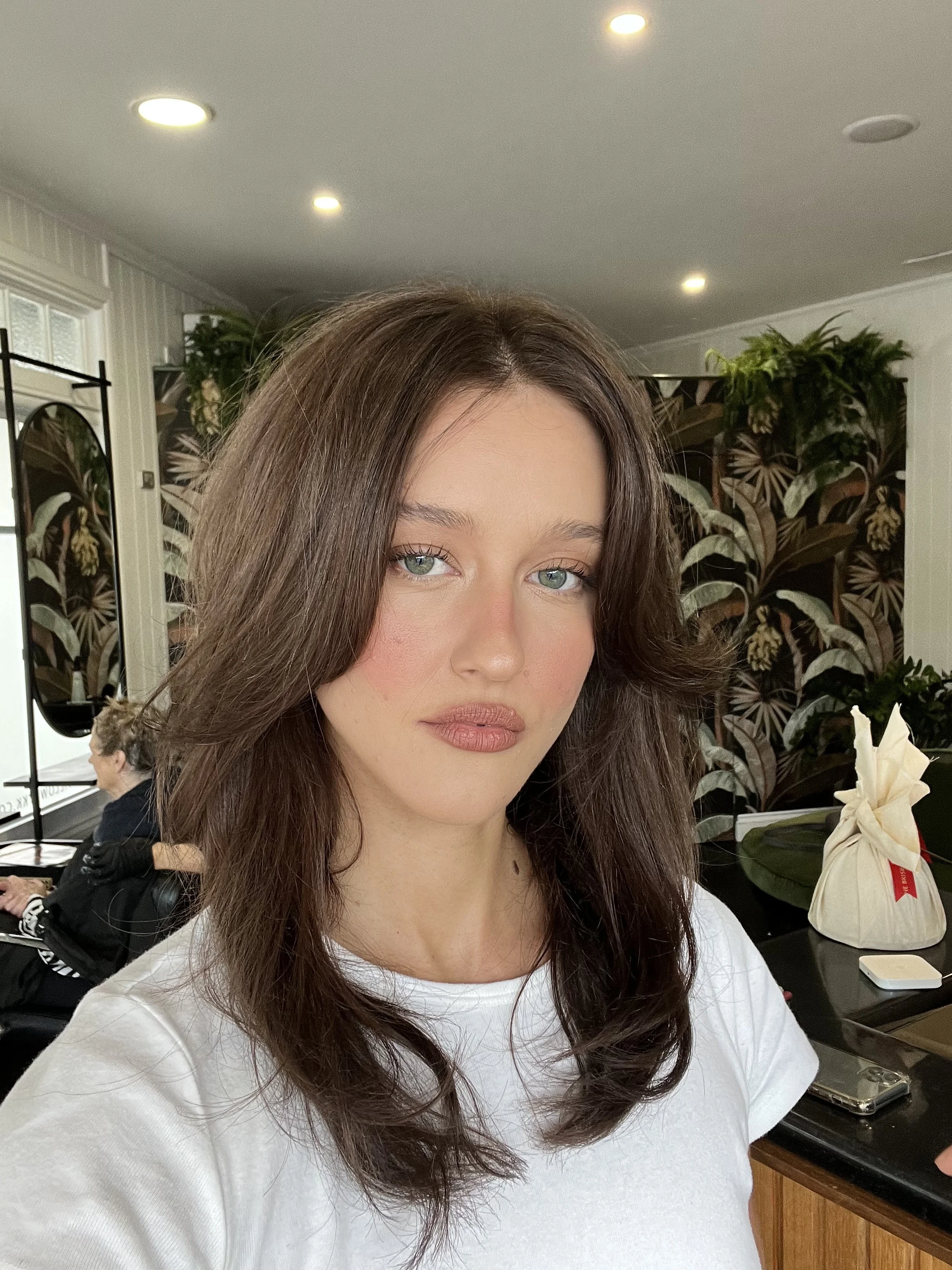 A woman with long, wavy brown hair and light-colored eyes taking a selfie in a room with floral wallpaper and plants in the background.