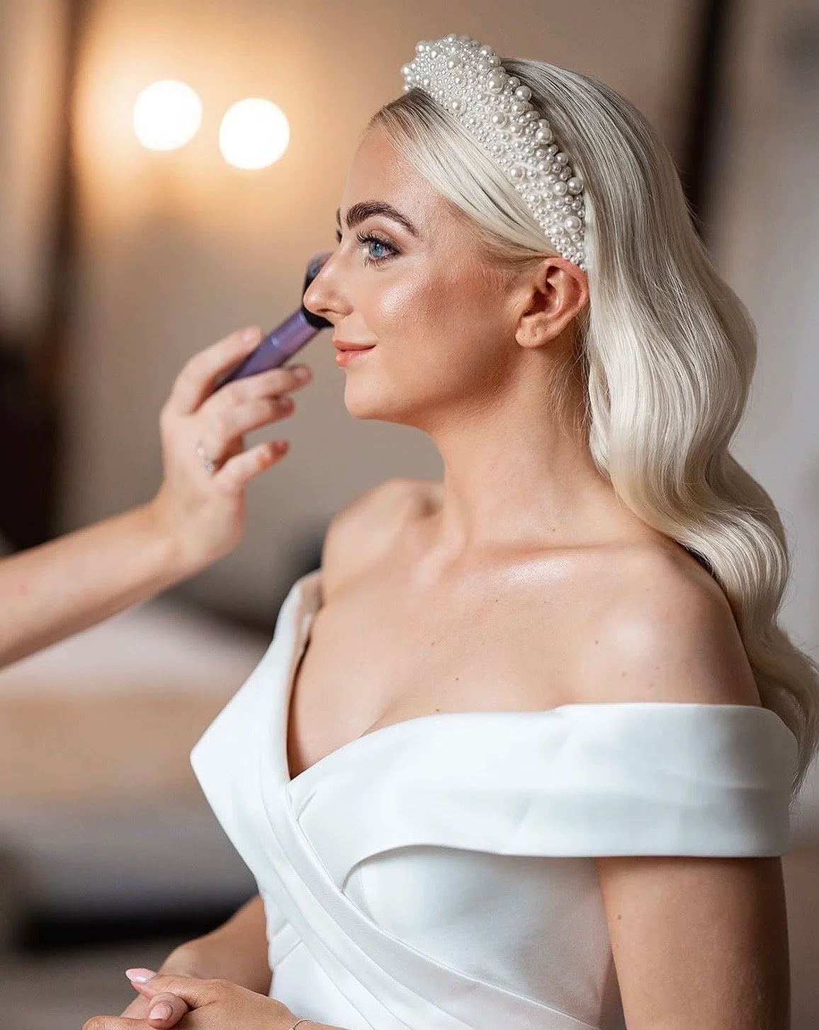 A woman with blonde hair wearing a pearl headband and a white off-shoulder dress is getting her makeup done, with a makeup brush near her face.