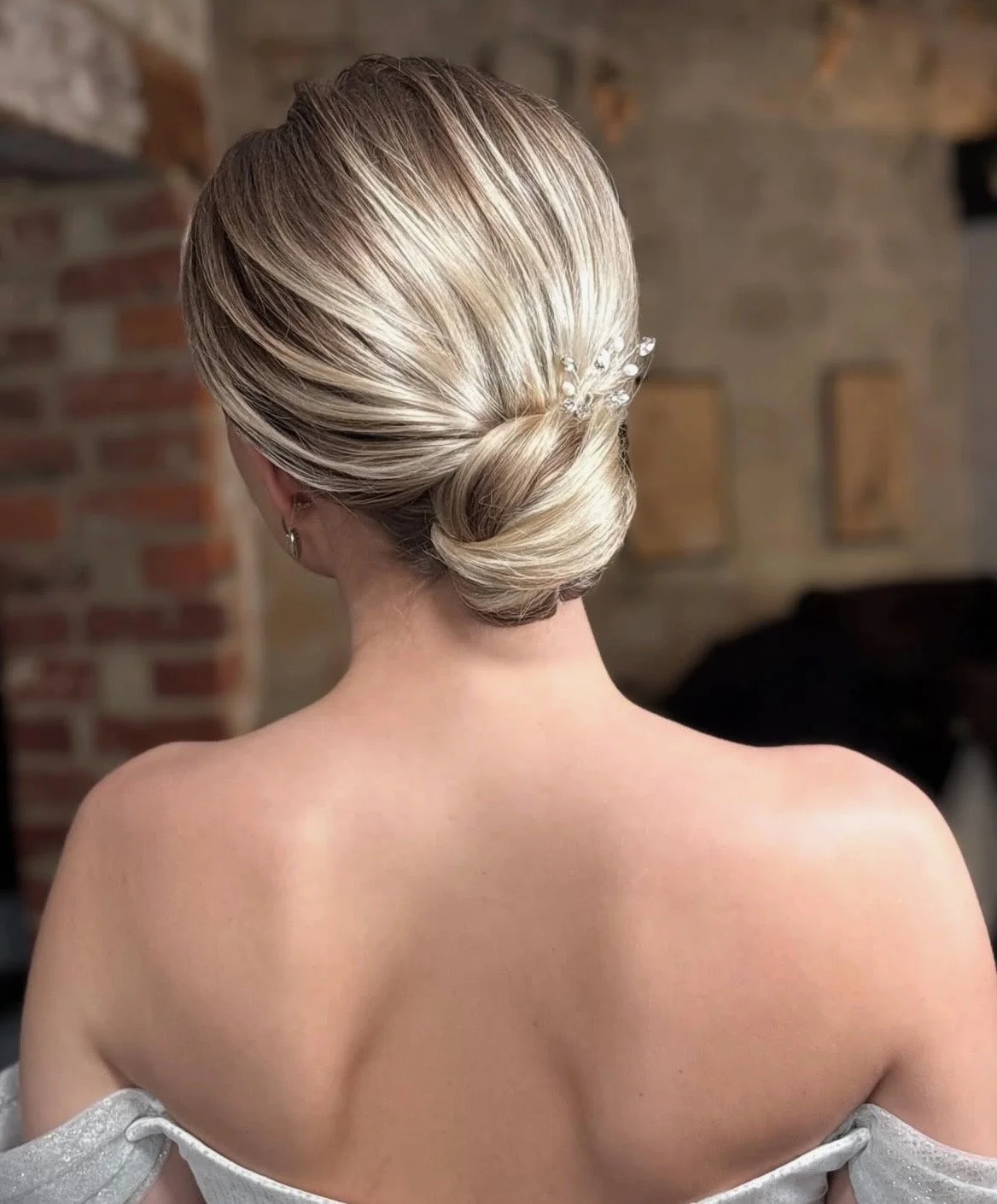 Back view of a woman with blonde hair in a low bun, decorated with a pearl and crystal hair accessory. She is topless, with an off-shoulder white garment, and a brick wall background.