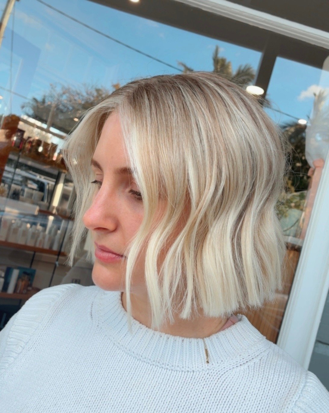 A woman with shoulder-length, wavy blonde hair and light skin, wearing a white sweater, looking down and outside a window on a clear day.