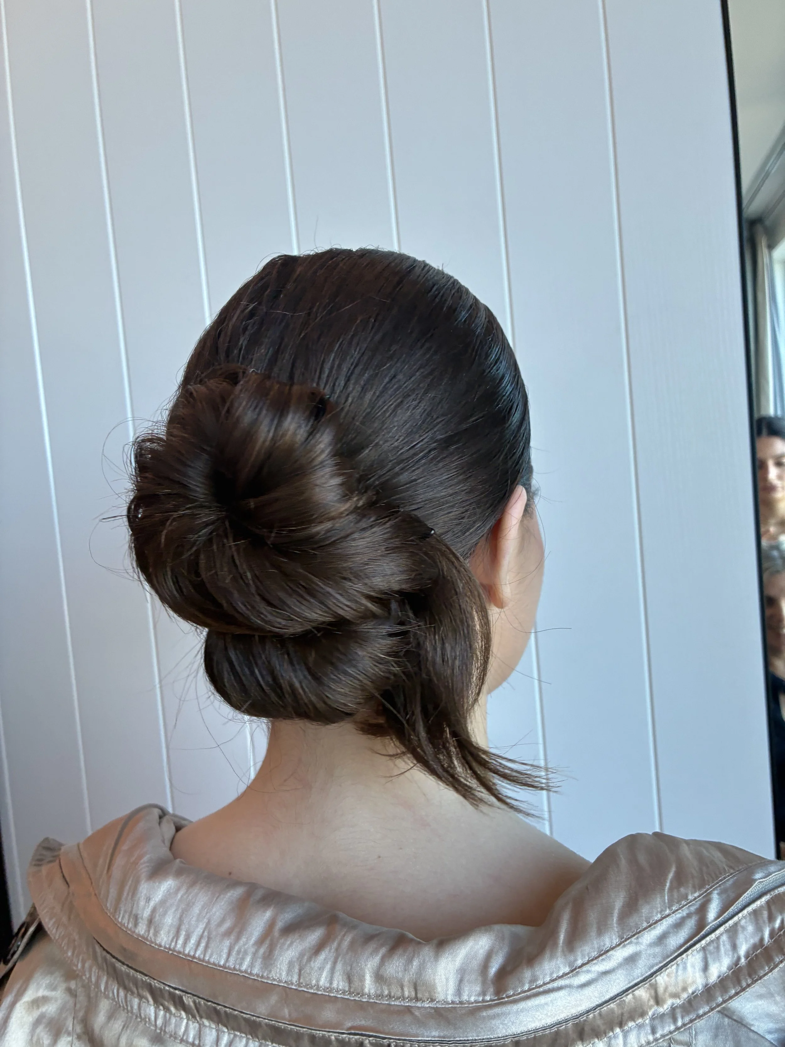 Back view of a woman with dark brown hair styled in an elegant bun, wearing a shiny bronze-colored jacket, standing in front of a light gray paneled wall, looking into a mirror, with a slight smile reflected in it.