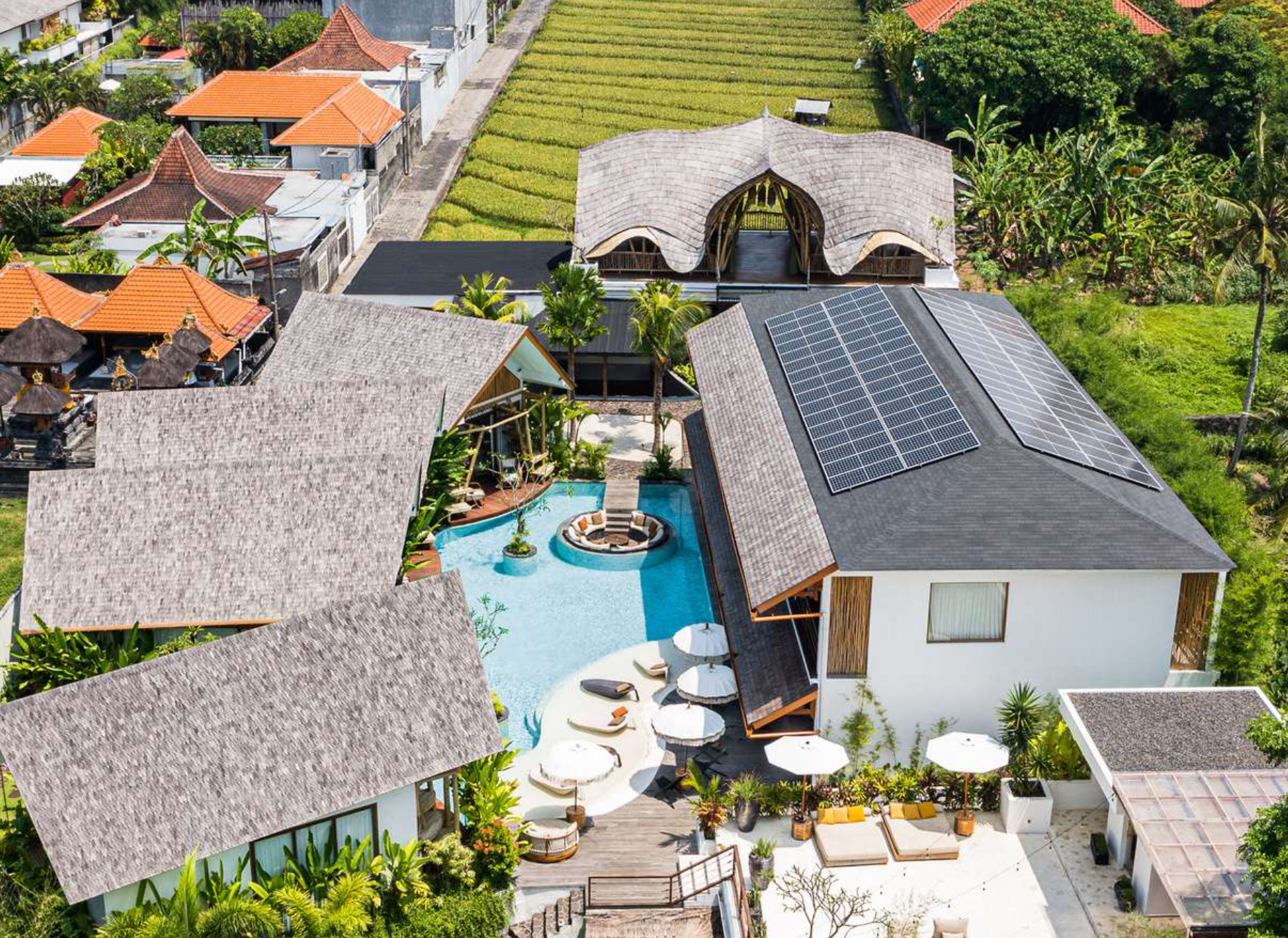 Aerial view of a luxurious villa with a swimming pool, surrounded by greenery and neighboring houses with red-tiled roofs, some of which have solar panels on their roofs.