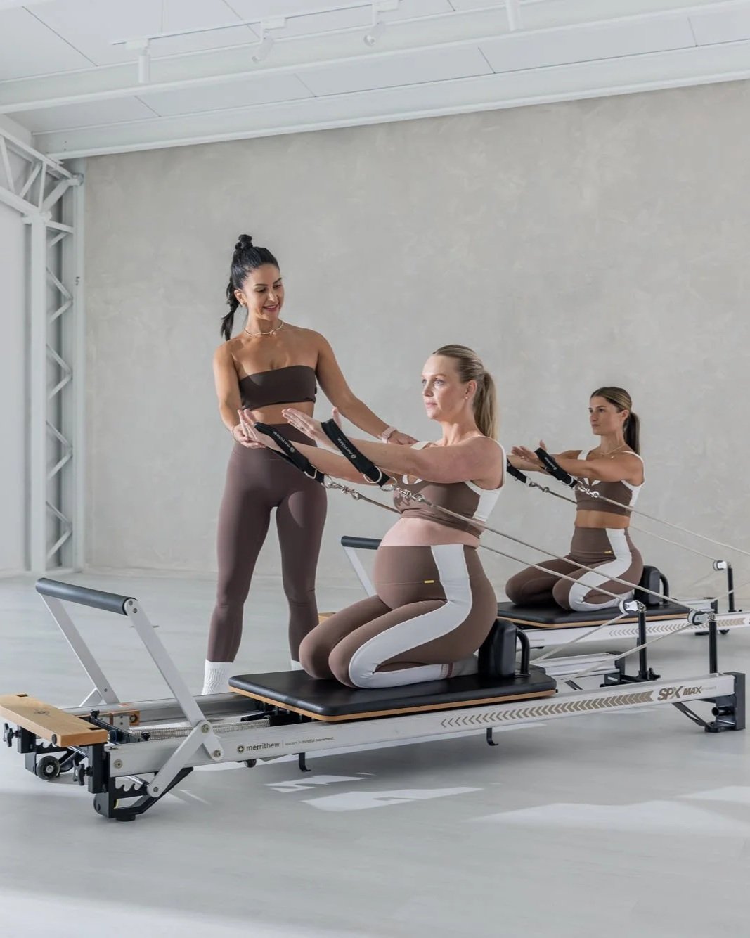 Woman practicing Pilates on a reformer machine in a bright room with sheer curtains and a potted plant.