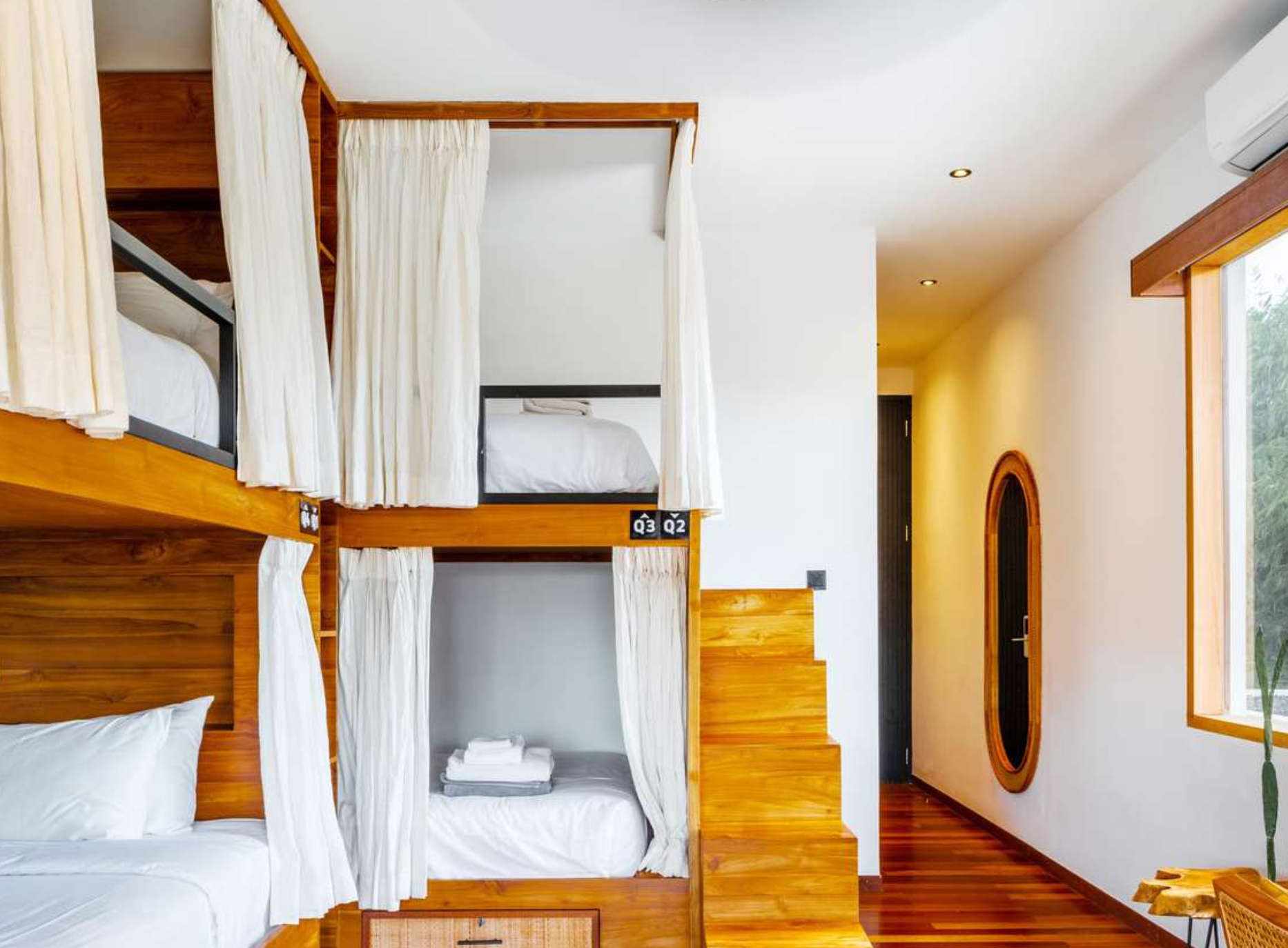 Interior of a cozy Airbnb with wooden bunk beds, white curtains, white linens, and a window with natural light.