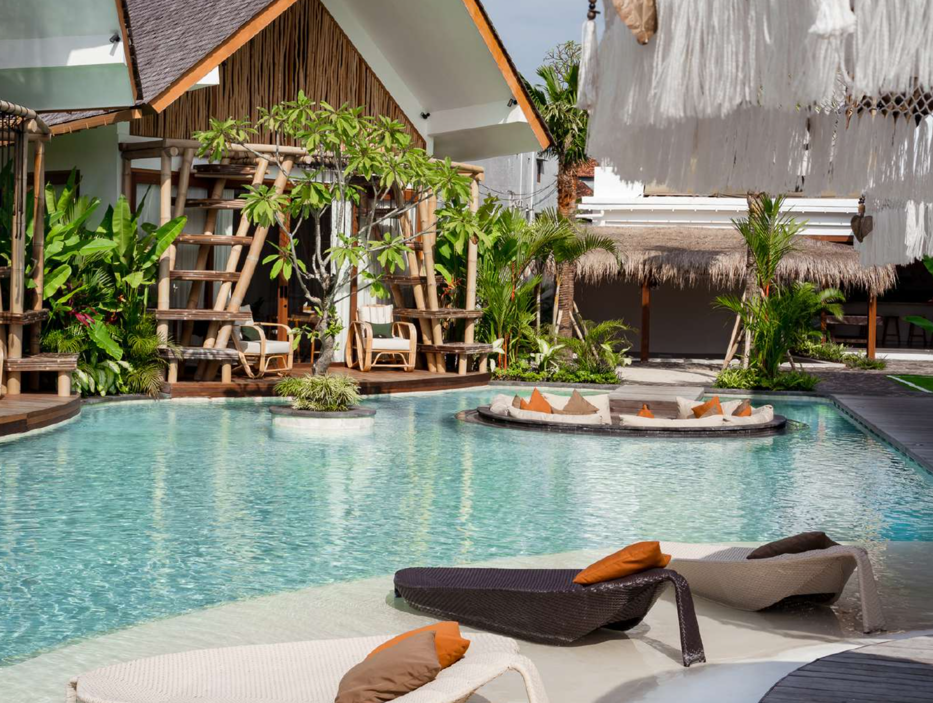 Luxury resort pool area with lounge chairs, tropical plants, bamboo structures, and thatched-roof buildings.