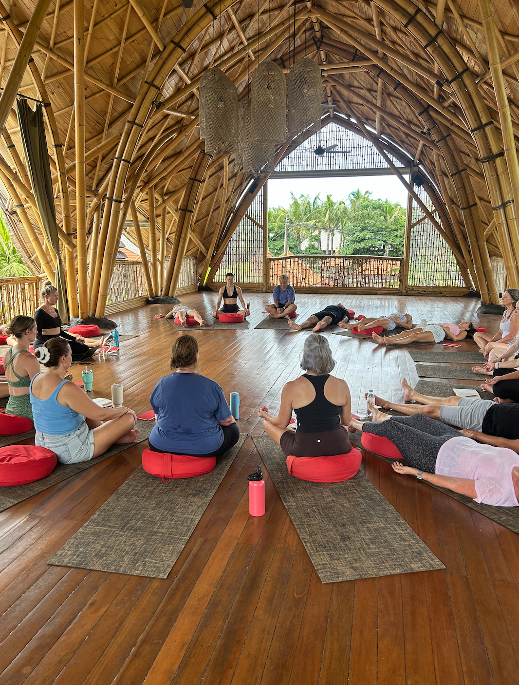 People participating in a yoga class inside a bamboo structure with a high vaulted ceiling, large windows, and wooden floors, overlooking a tropical landscape.