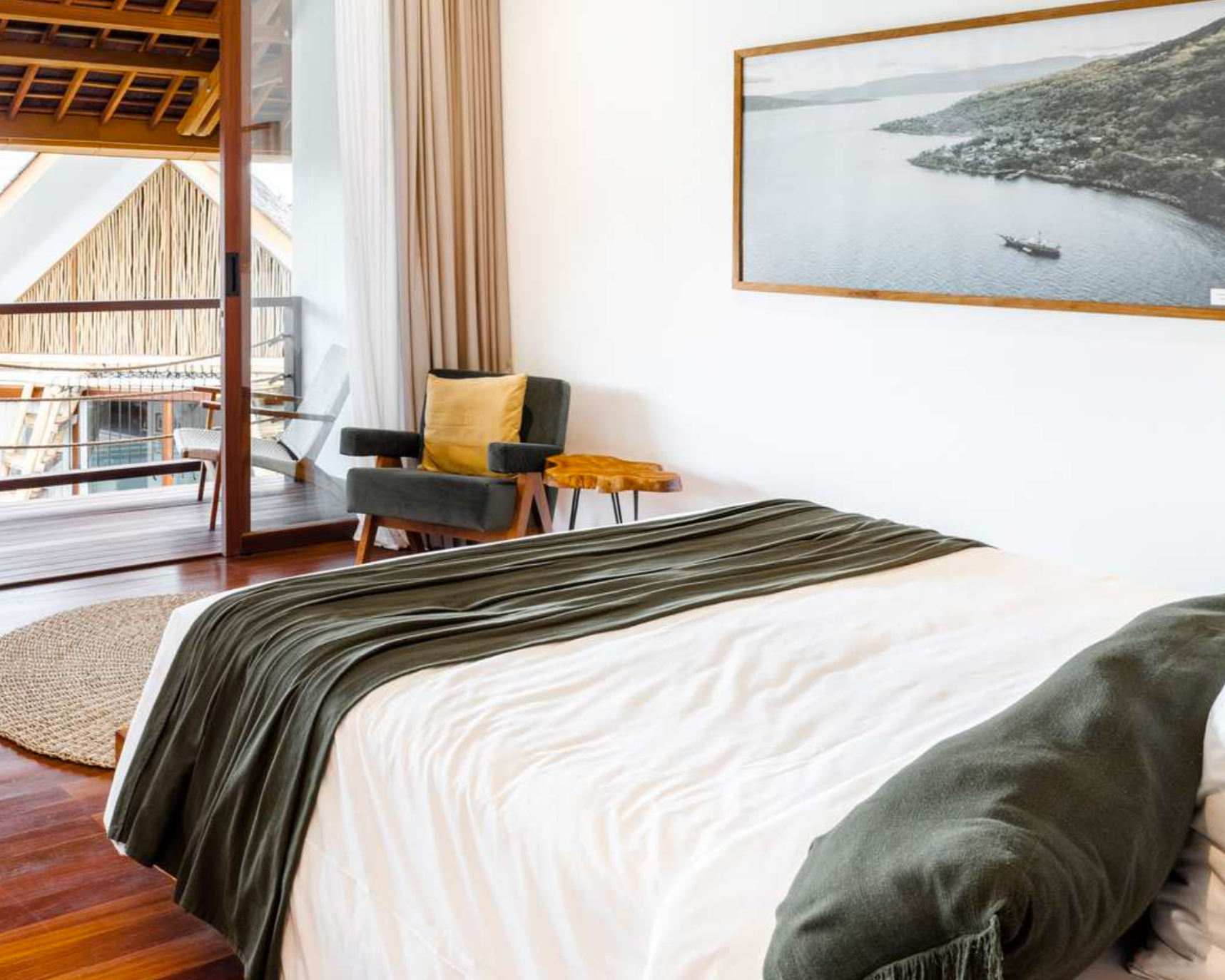 A bedroom with a bed, a chair with a pillow, and a small side table. There is a large picture of a river and shoreline on the wall. A sliding glass door leads to a balcony with outdoor seating.