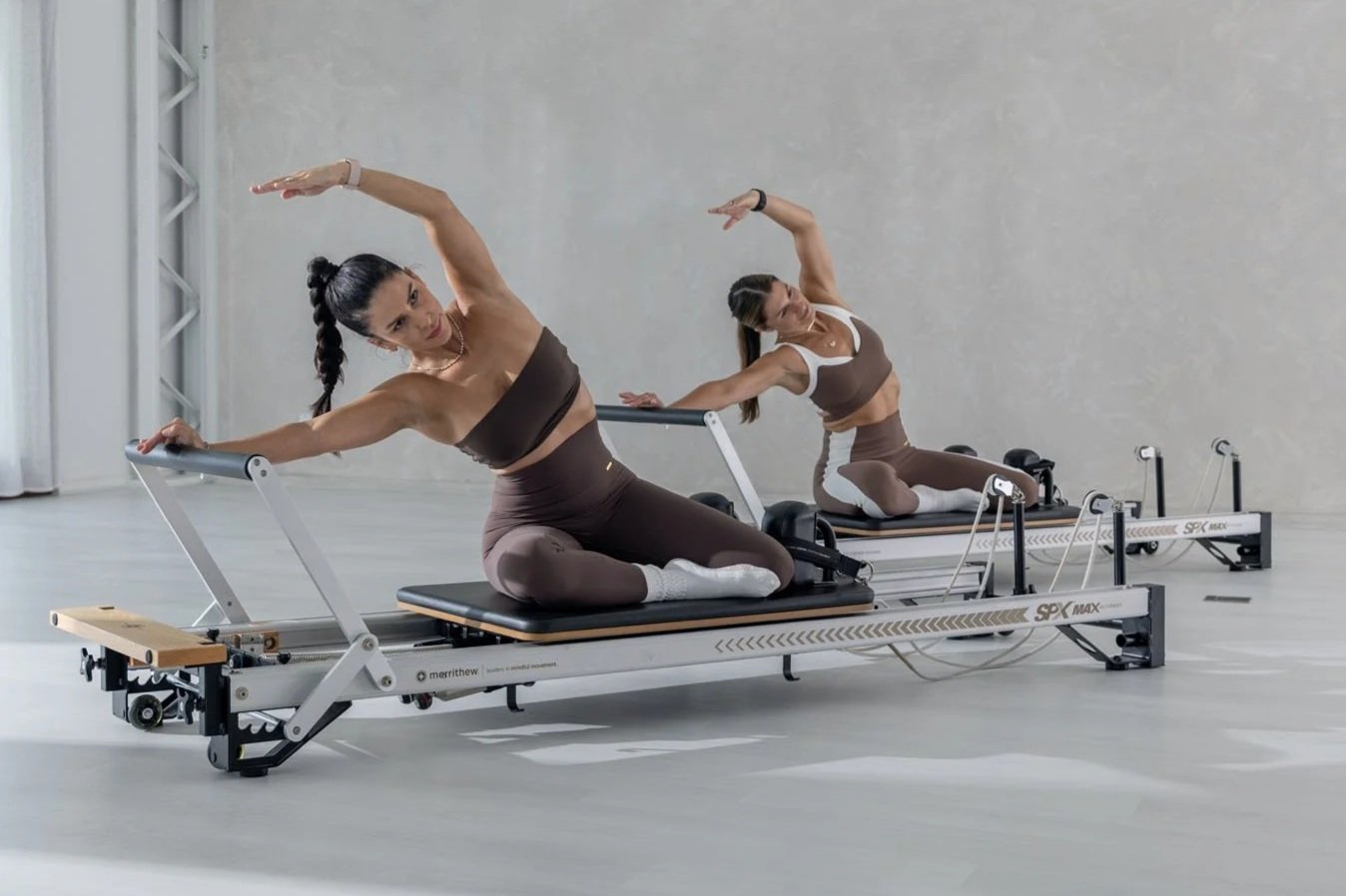 Woman in workout clothes doing a Pilates exercise on a reformer machine in a bright room with white curtains and a large potted plant.