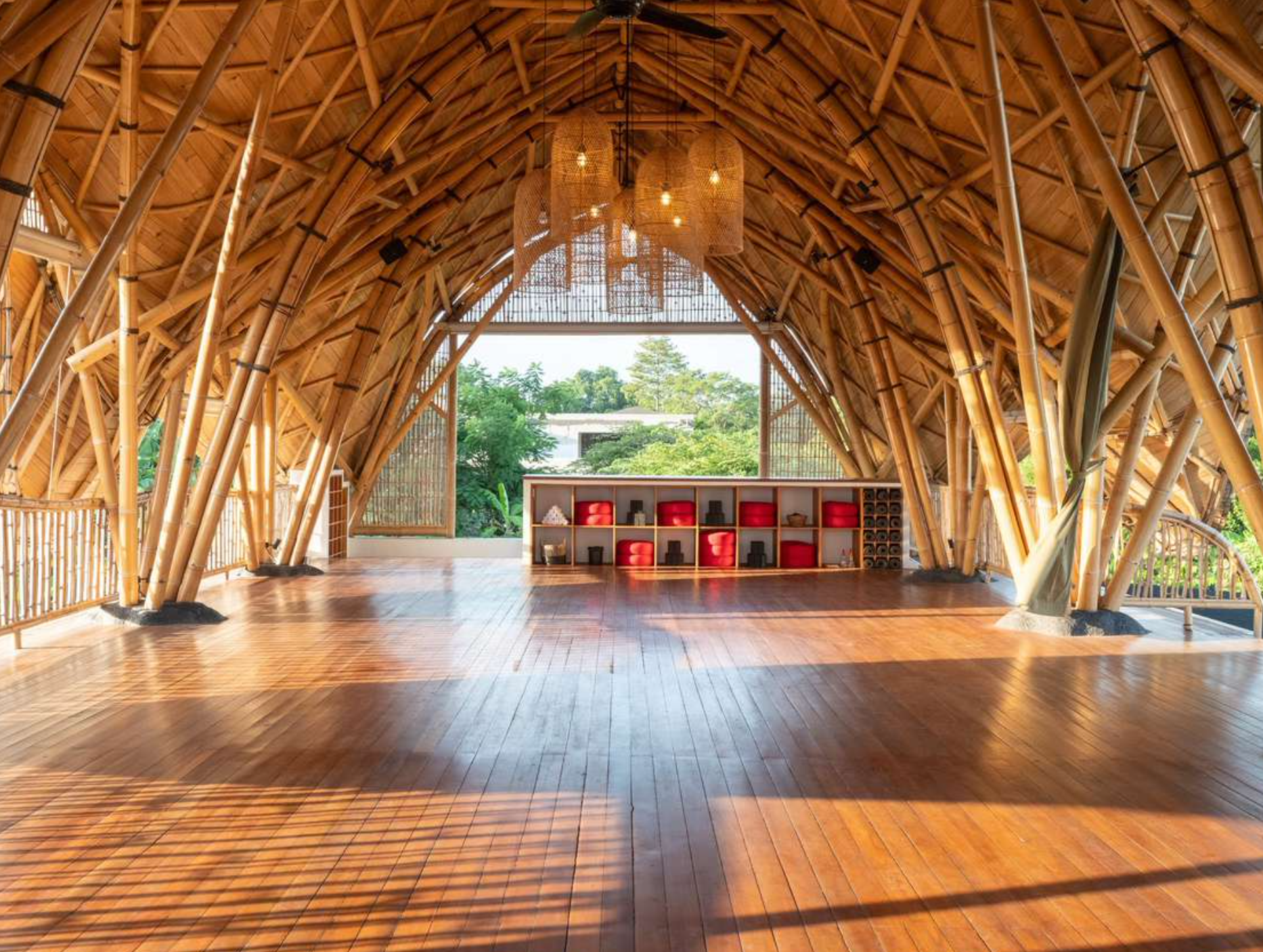 Interior of a bamboo structure with a high arched ceiling, hardwood floor, and large open window showing green trees outside. There is a red and white shelving unit with meditation cushions and decorative items.