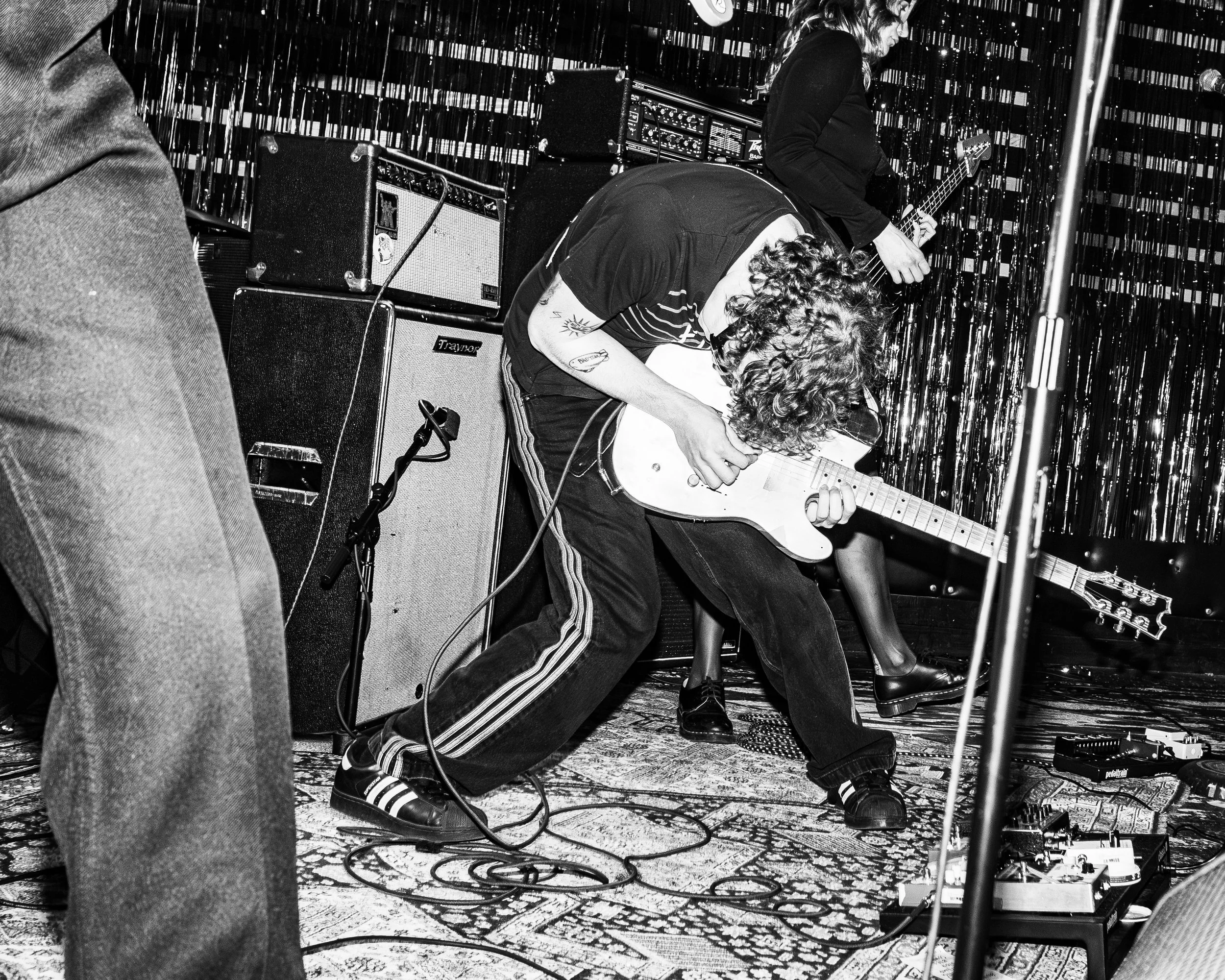 A musician with curly hair, wearing a striped t-shirt and tracksuit pants, is performing on stage with an electric guitar, surrounded by amplifiers and equipment, with another guitarist in the background, in front of a shiny curtain.