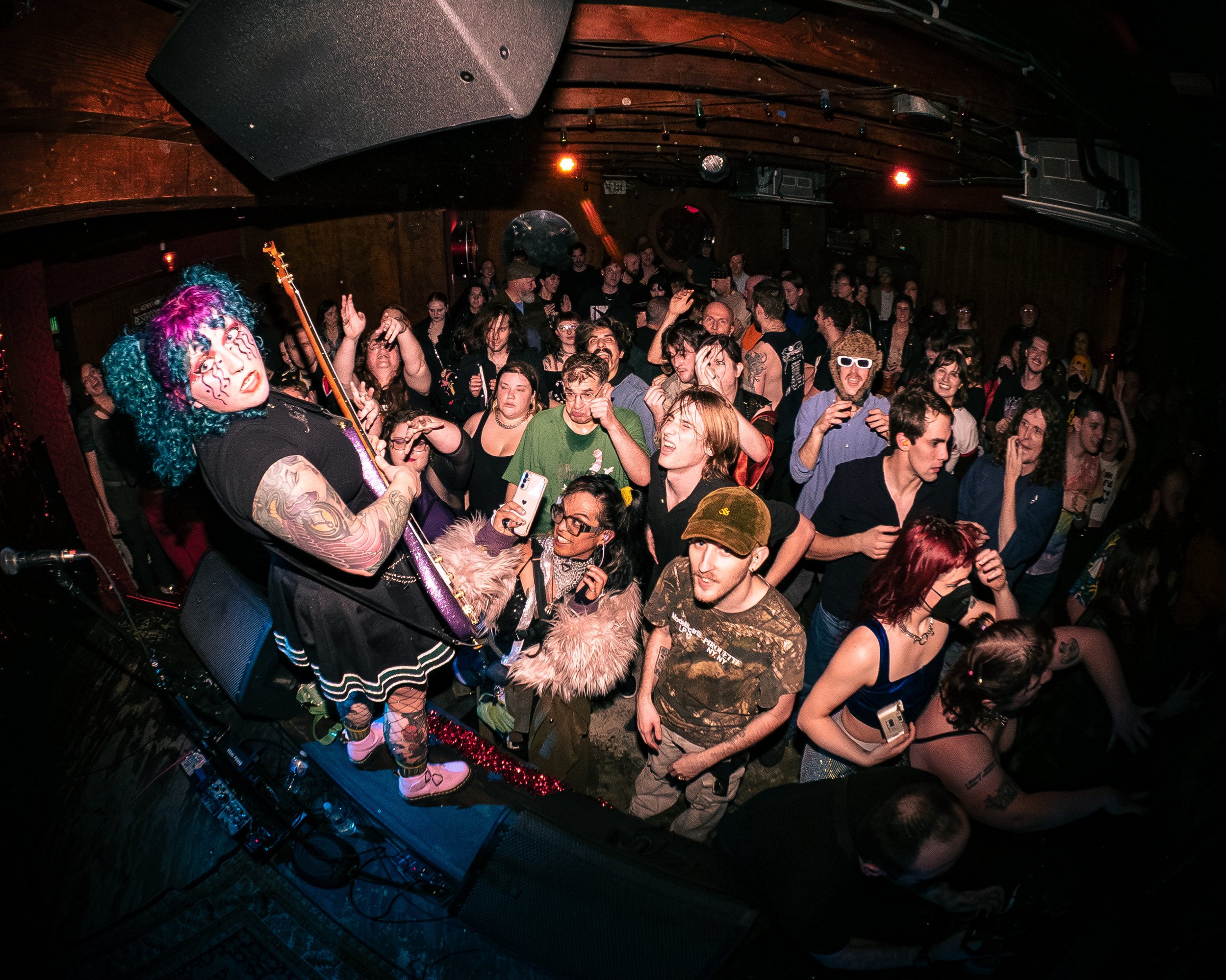 A lively crowd of people at a Zookraught concert, dancing and enjoying music, with Stephanie Jones on stage holding a guitar, dressed in colorful, eccentric clothing and vibrant, multicolored hair.