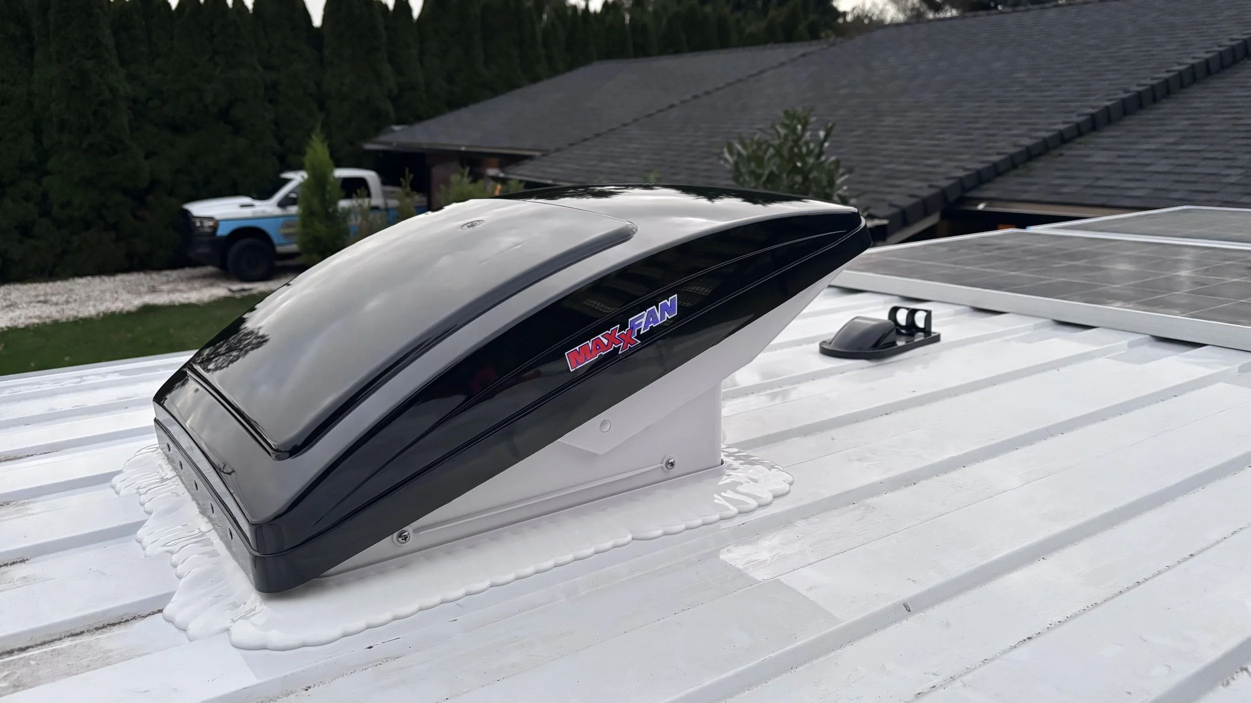 A MAXXFAN roof vent installed on a white metal roof, with a solar panel nearby and a background of trees and a house.