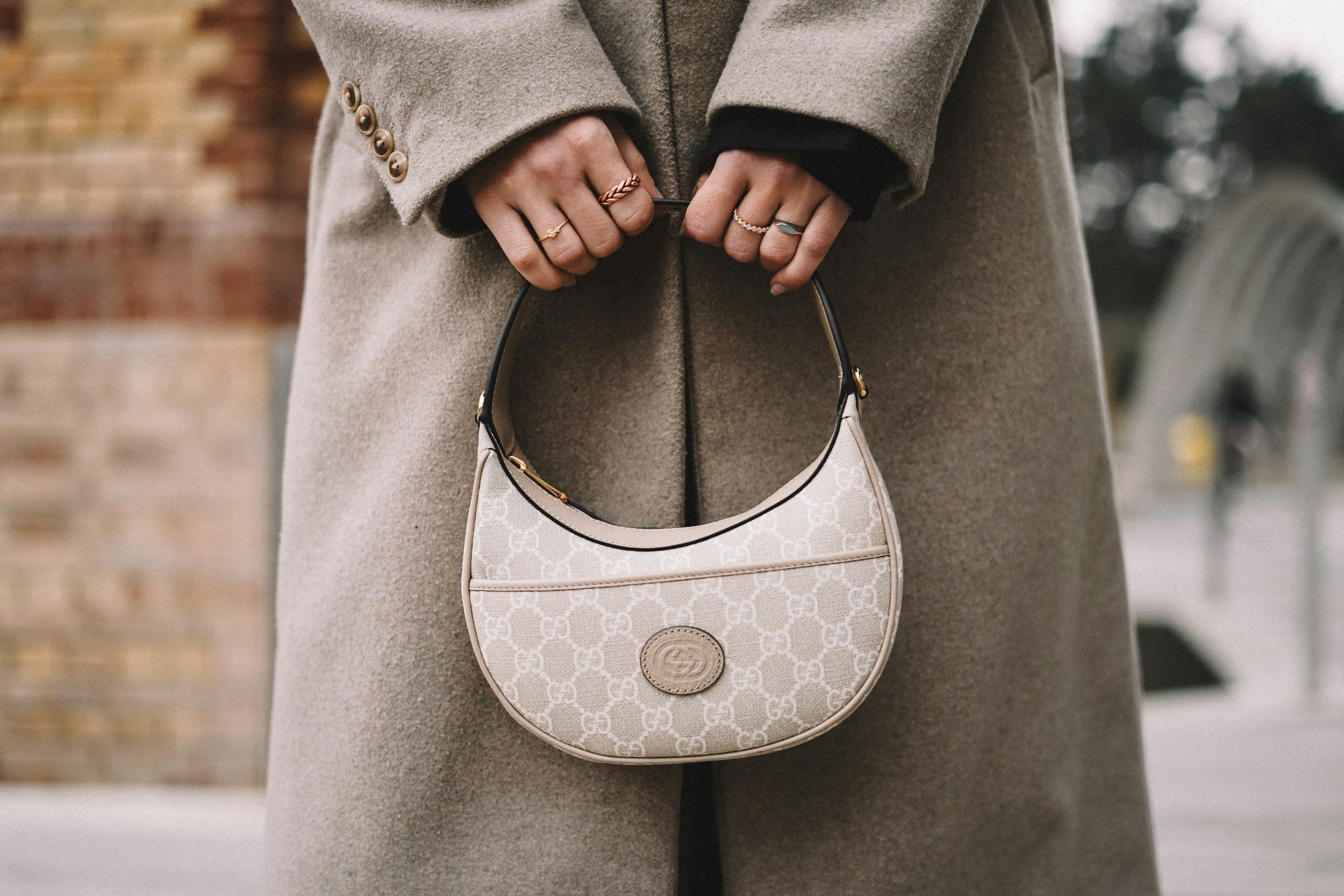 Person holding a small beige designer handbag with a pattern and logo, wearing multiple rings, dressed in a beige coat with button details, outdoors with blurred background.