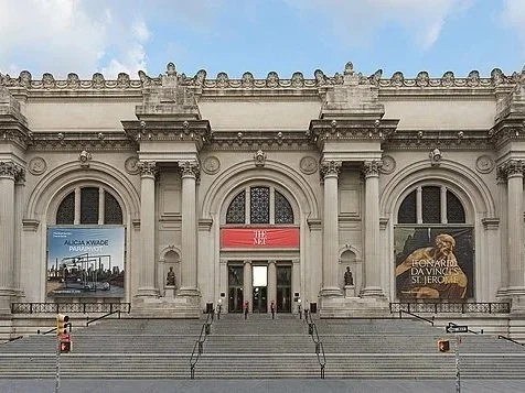 The image shows the front facade of a grand, classical-style building with large arched windows and ornate architectural details. There are steps leading up to the entrance and two large banners on either side of the building.