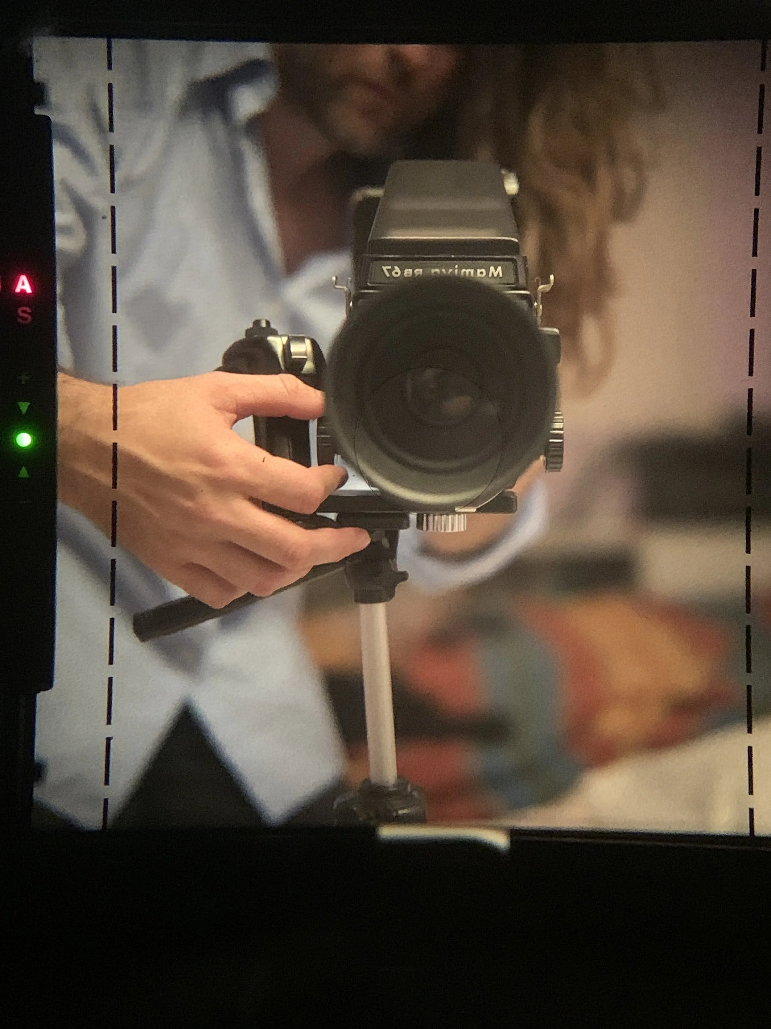 Photographer taking a mirror selfie with a professional camera, visible hand holding the camera, and a person with long curly hair in the background.