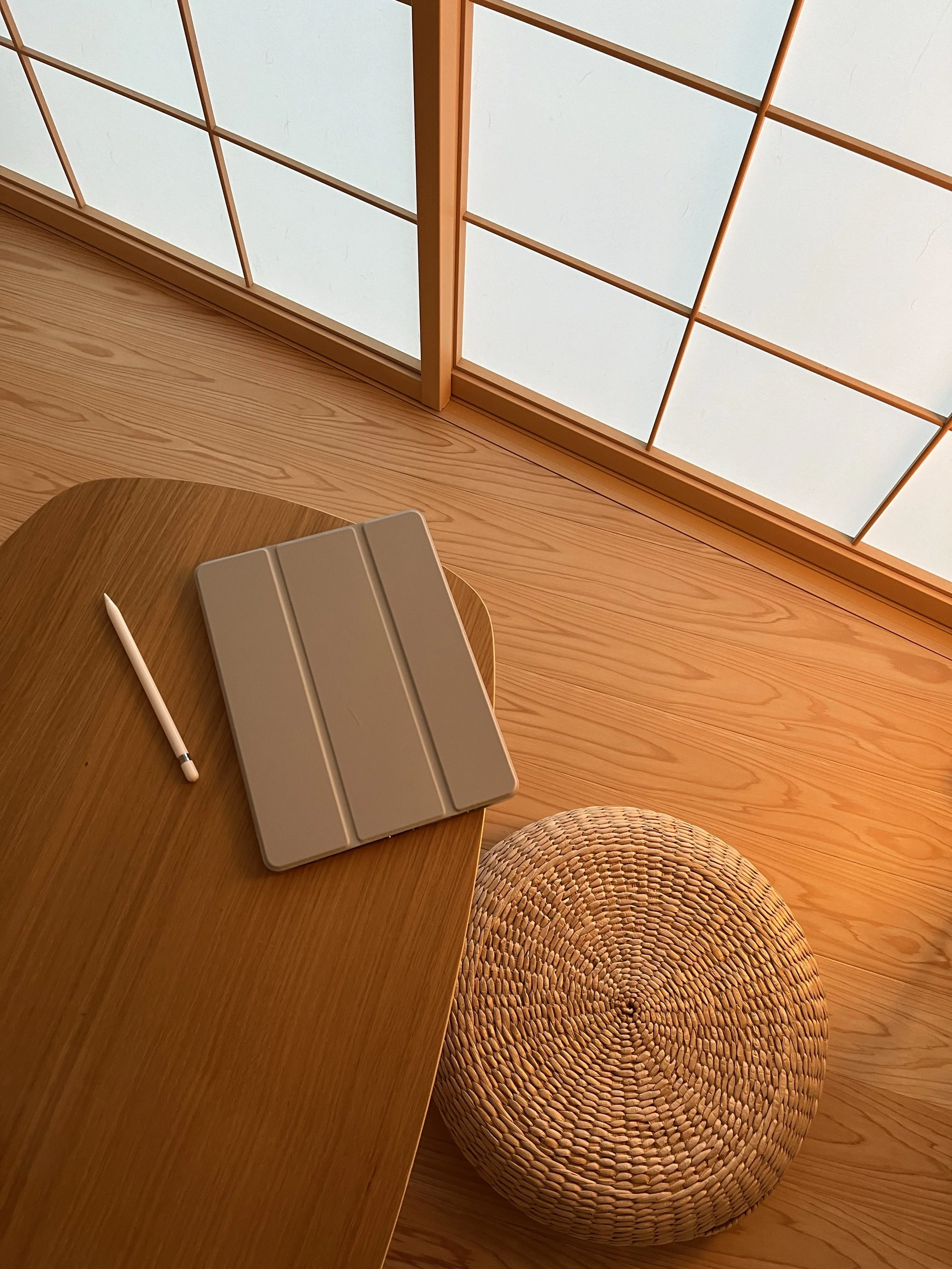 A wooden table with a gray tablet and a white stylus on it, next to a woven round cushion, near shoji-style windows with paper panels.