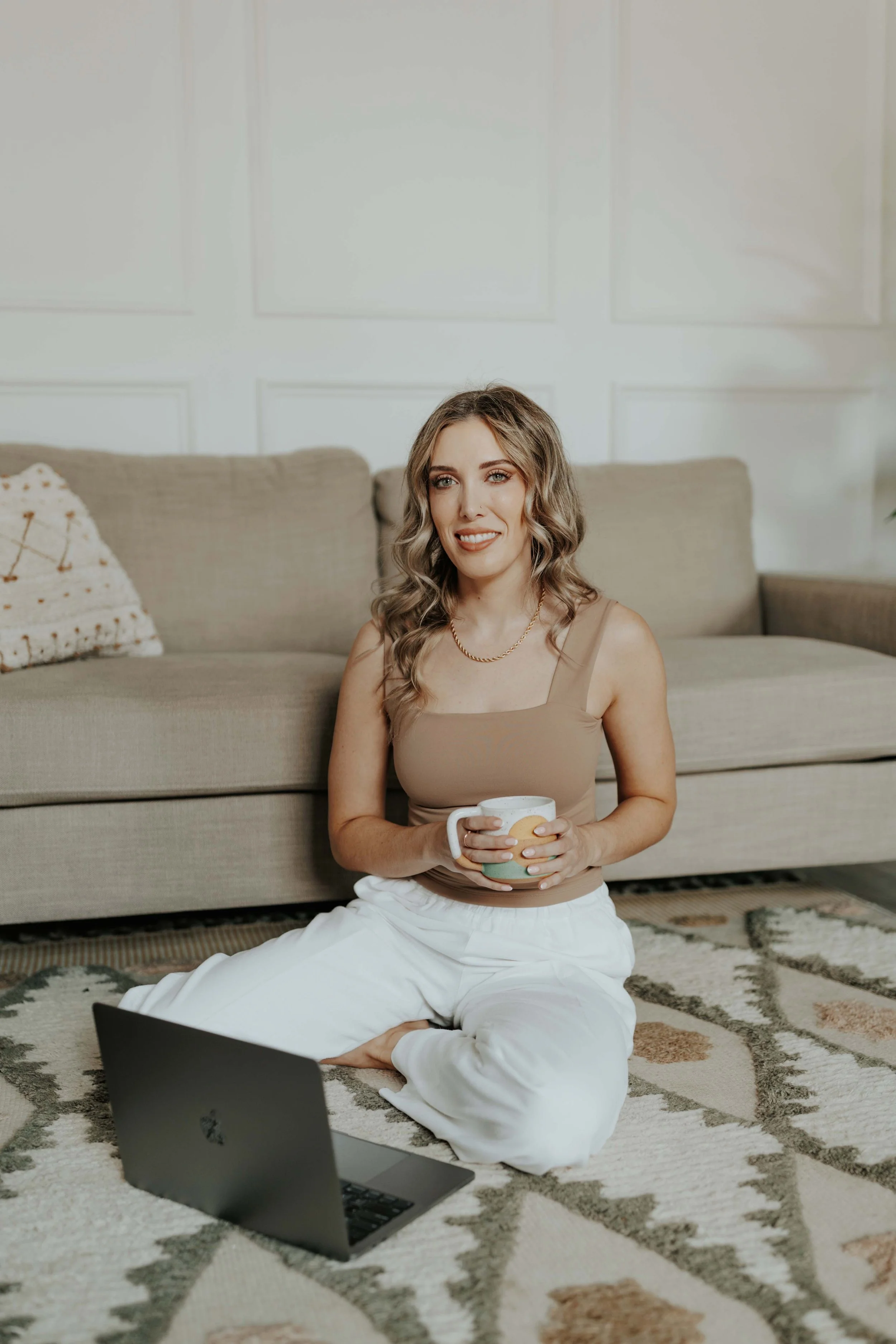 A woman with wavy blonde hair sitting on a patterned rug on the floor, holding a mug in her hands, with a laptop open in front of her, sitting in a living room with a beige sofa and a decorative pillow.
