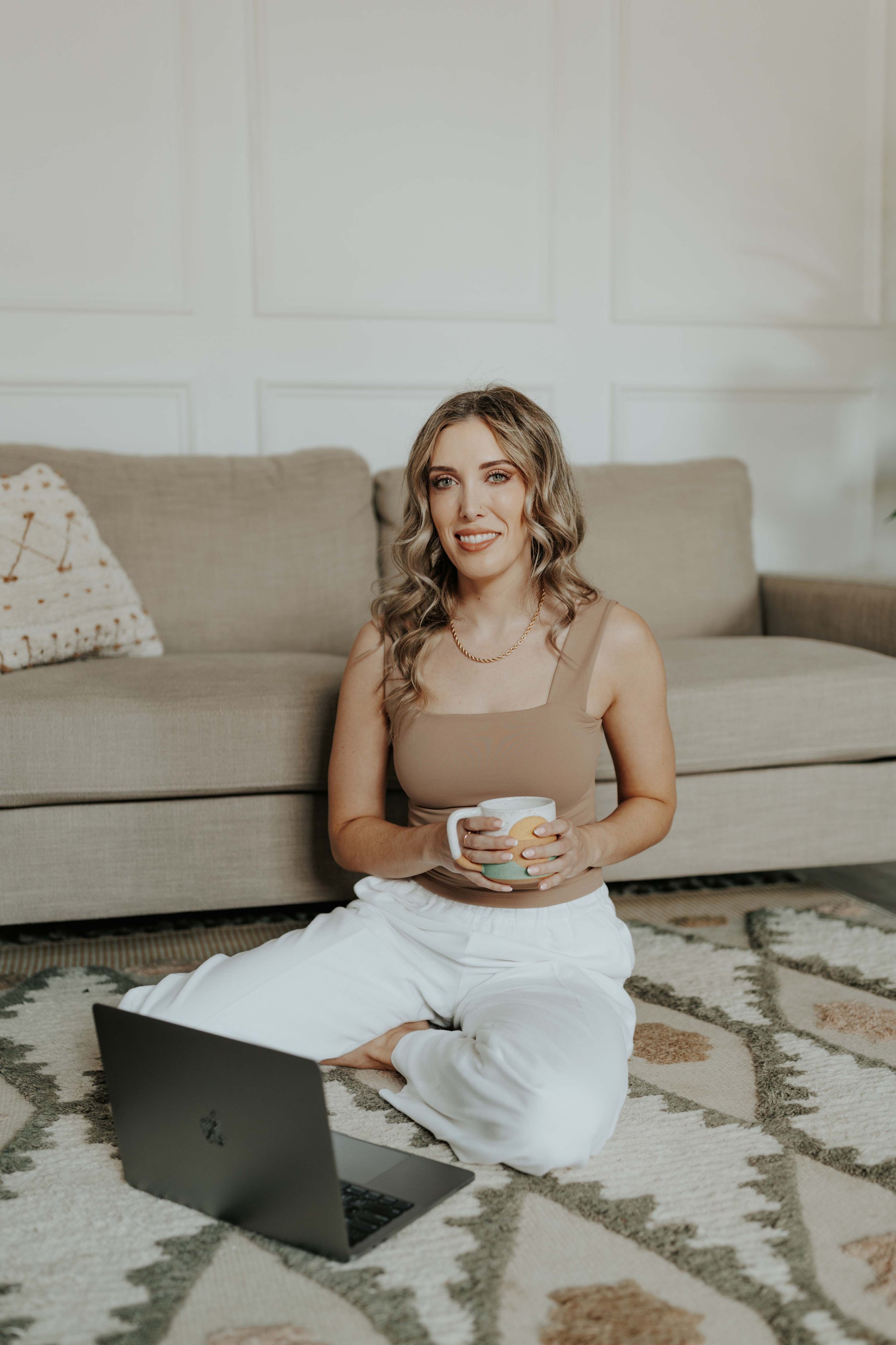 Photo of Becca Gregory, LICSW, sitting cross-legged on a patterned rug with a mug and laptop in a cozy living room.
