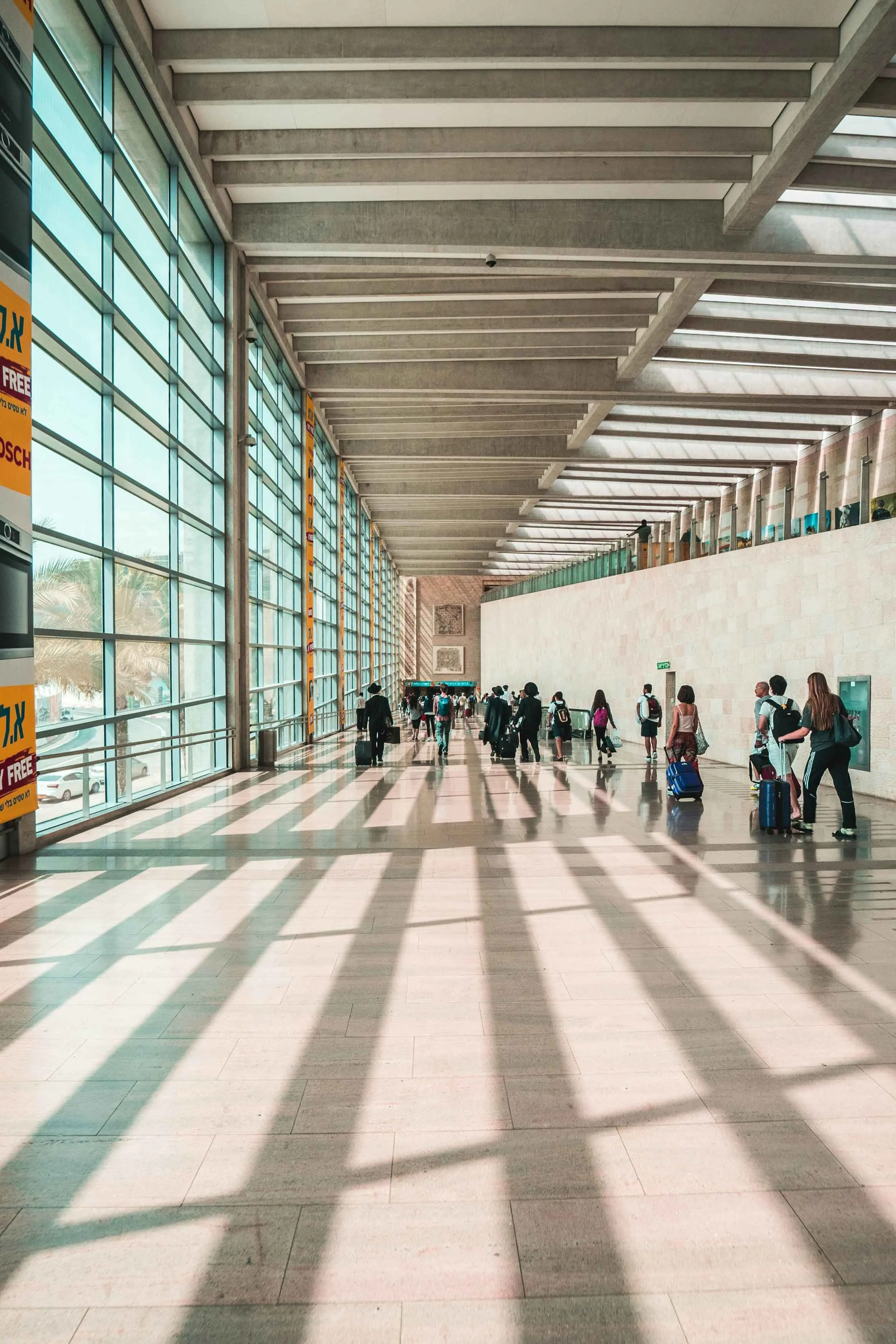 An airport hall with many people walking towards the entrance or exit of the hall, carrying suitcases