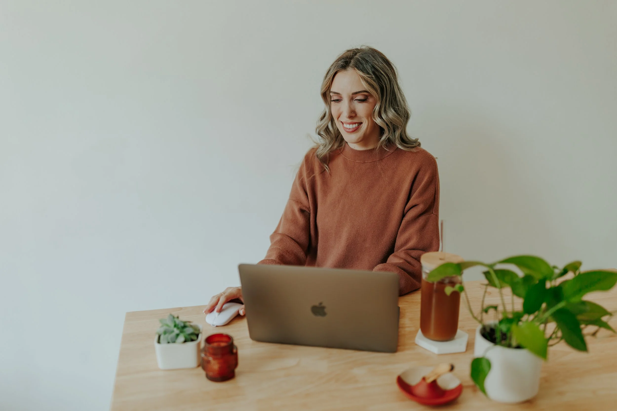 A young woman with wavy blonde hair sitting at a wooden table, working on a silver MacBook. The table has various potted plants, a jar with a candle, and a decorative bowl. She is wearing a brown sweater and smiling while using a computer mouse.