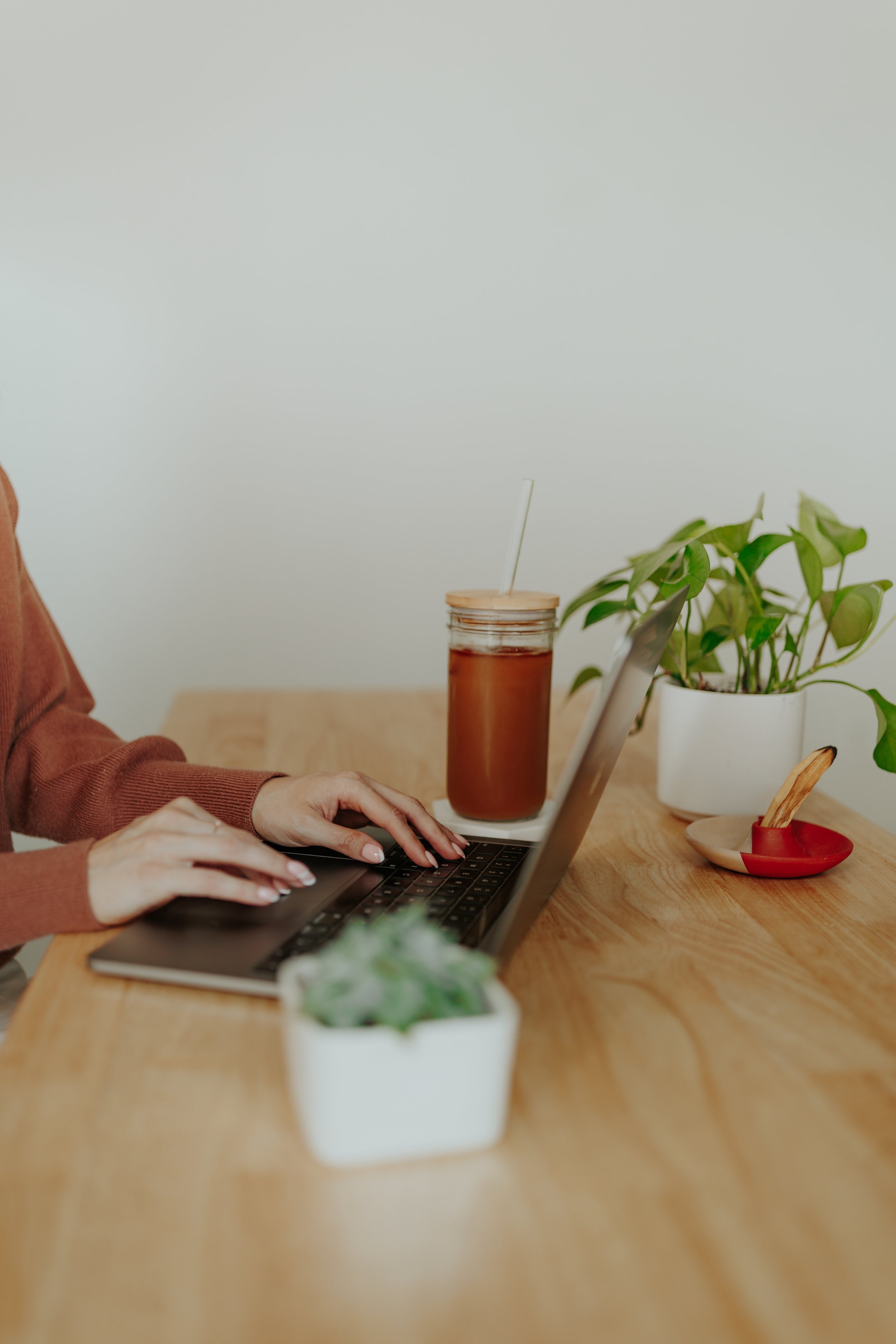 Person working on a laptop at a wooden desk with potted plants, a jar of tea, and a red bird-shaped dish.