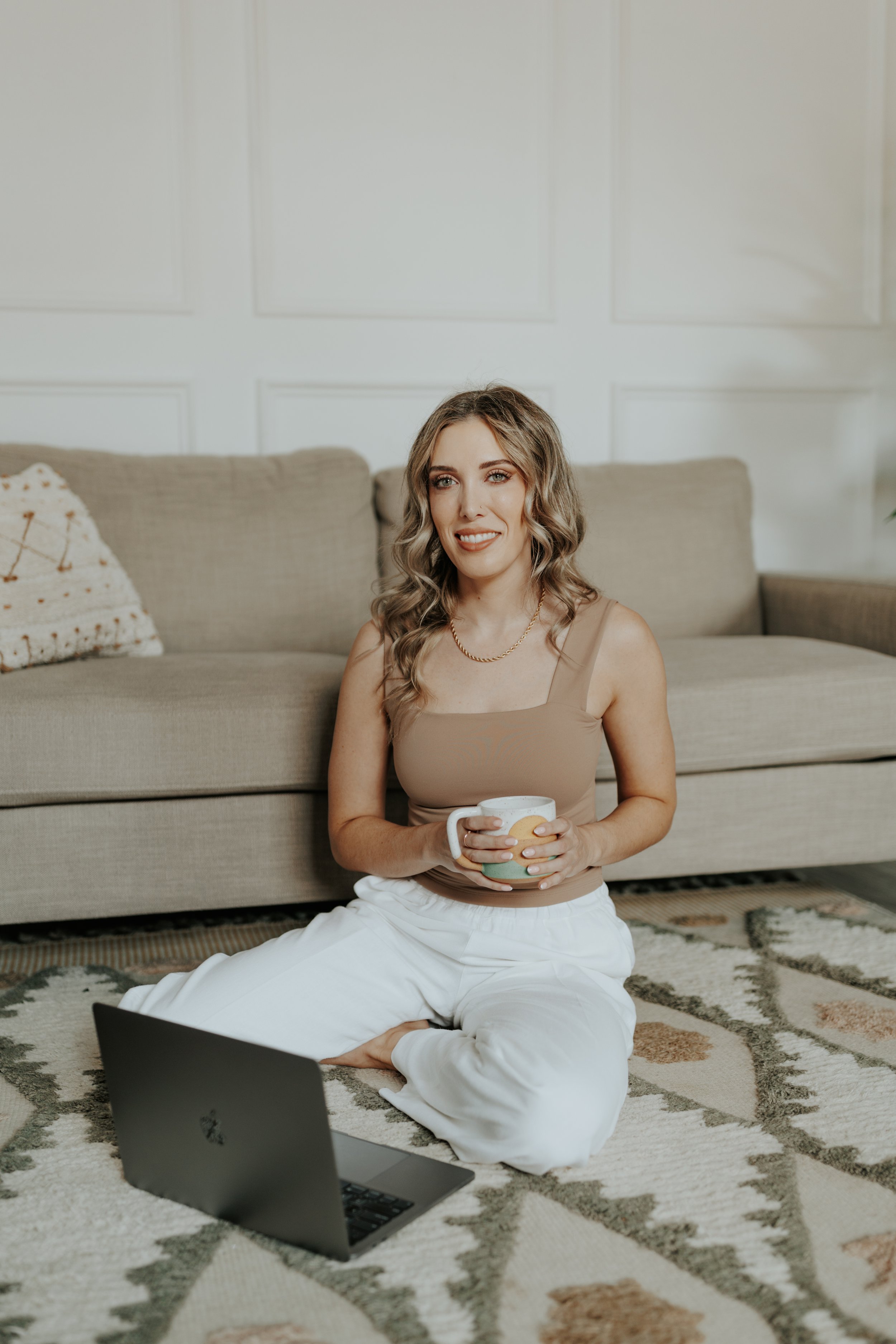 Photo of Becca Gregory, LICSW, sitting cross-legged on a patterned rug with a mug and laptop in a cozy living room.