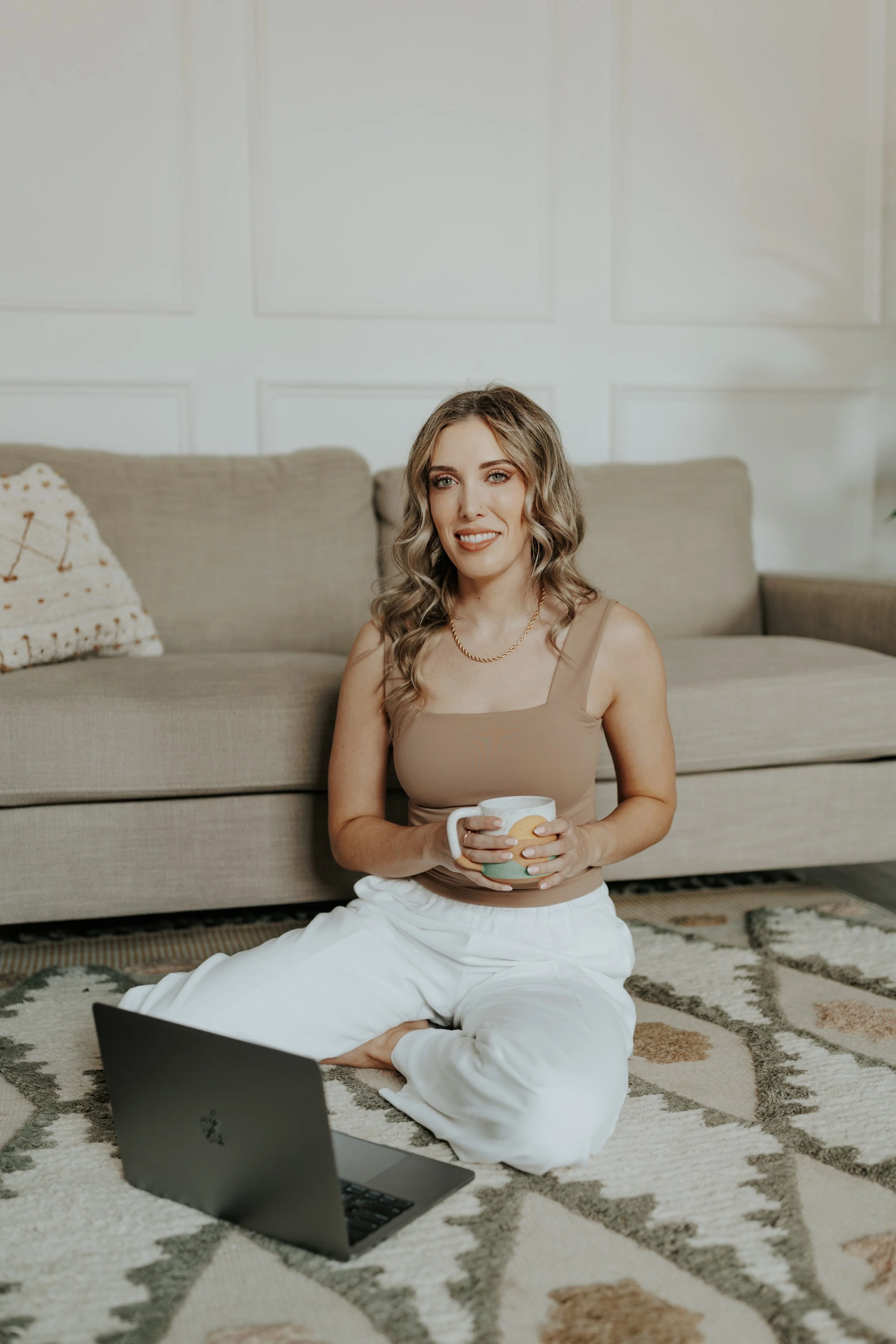 A woman with wavy blonde hair sitting on a patterned rug on the floor, holding a mug in her hands, with a laptop open in front of her, sitting in a living room with a beige sofa and a decorative pillow.
