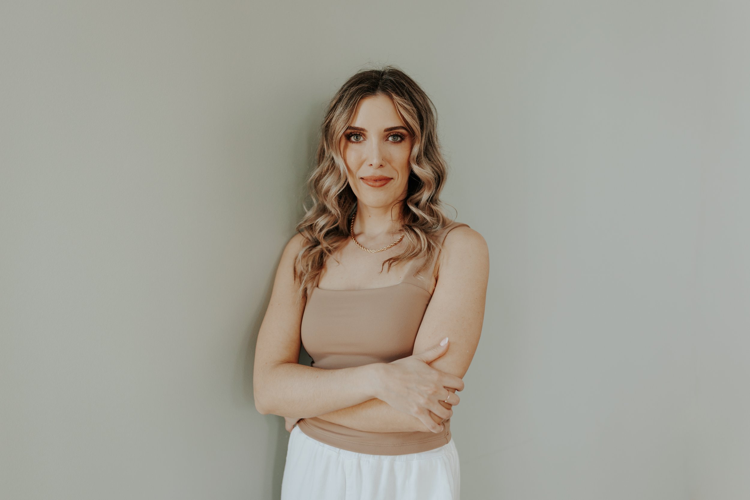 A woman with wavy blonde hair, wearing a beige sleeveless top, gold necklace, and white pants, standing against a plain light-colored wall.