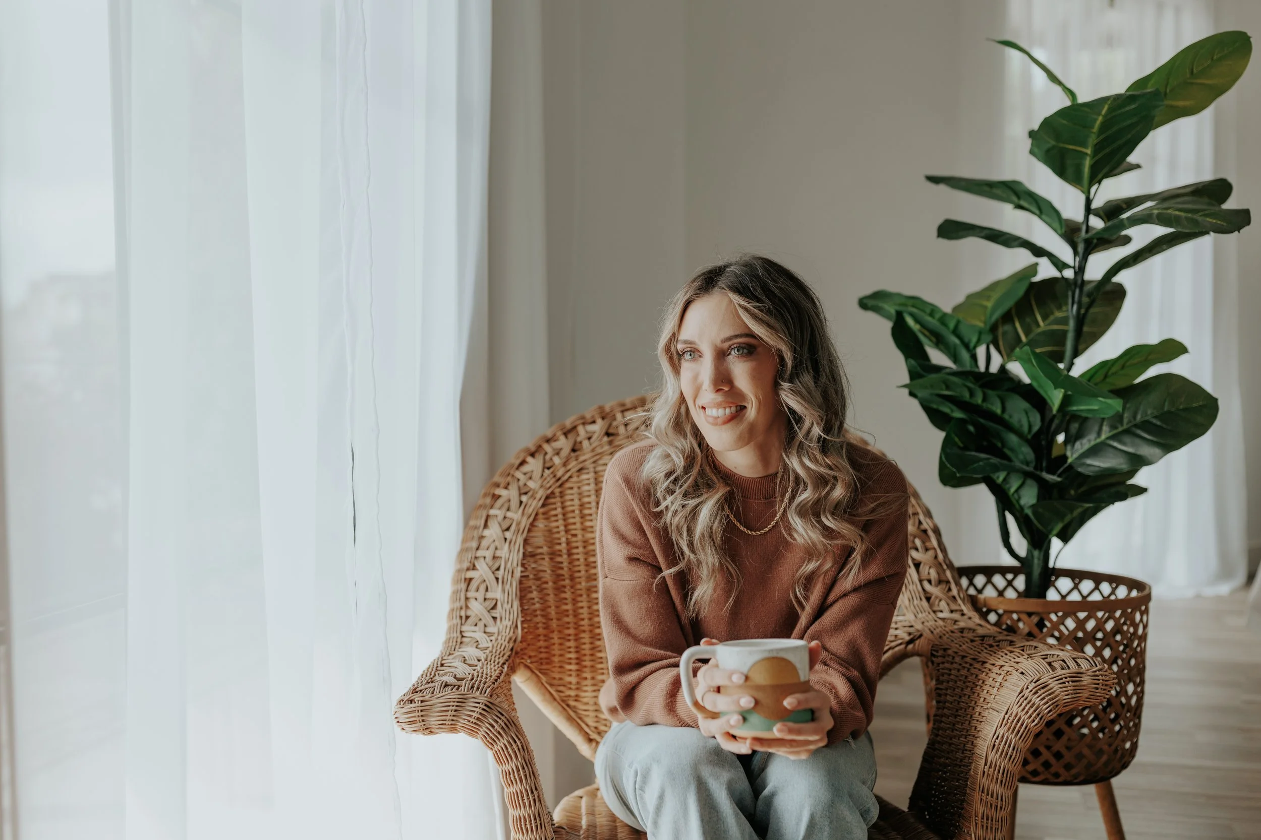 Photo of Becca Gregory, LICSW sitting in a wicker chair with a mug, smiling beside a large green plant in soft natural light.