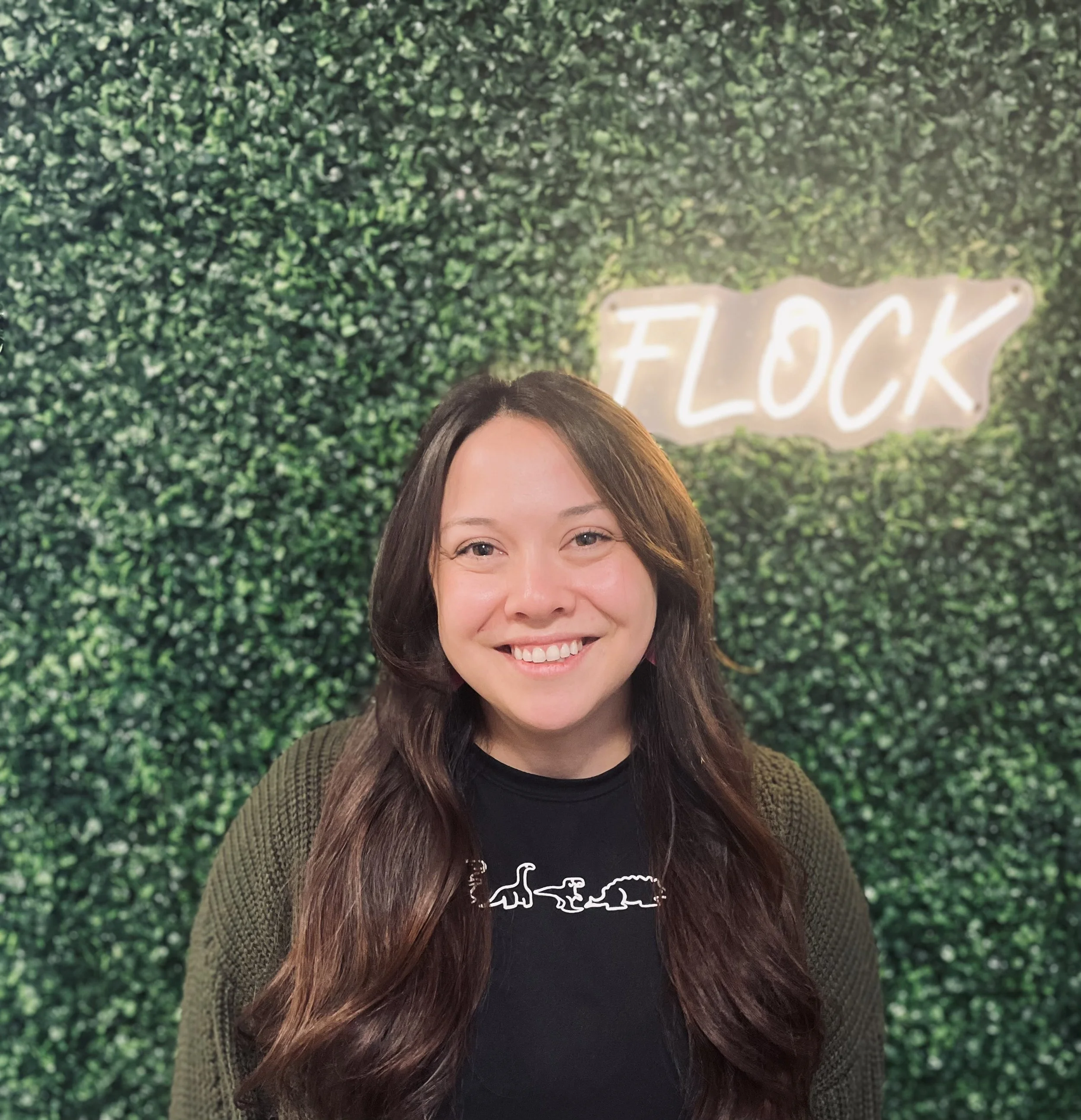 A smiling woman with long dark hair standing in front of a green foliage wall with a illuminated sign that reads 'FLOCK'.