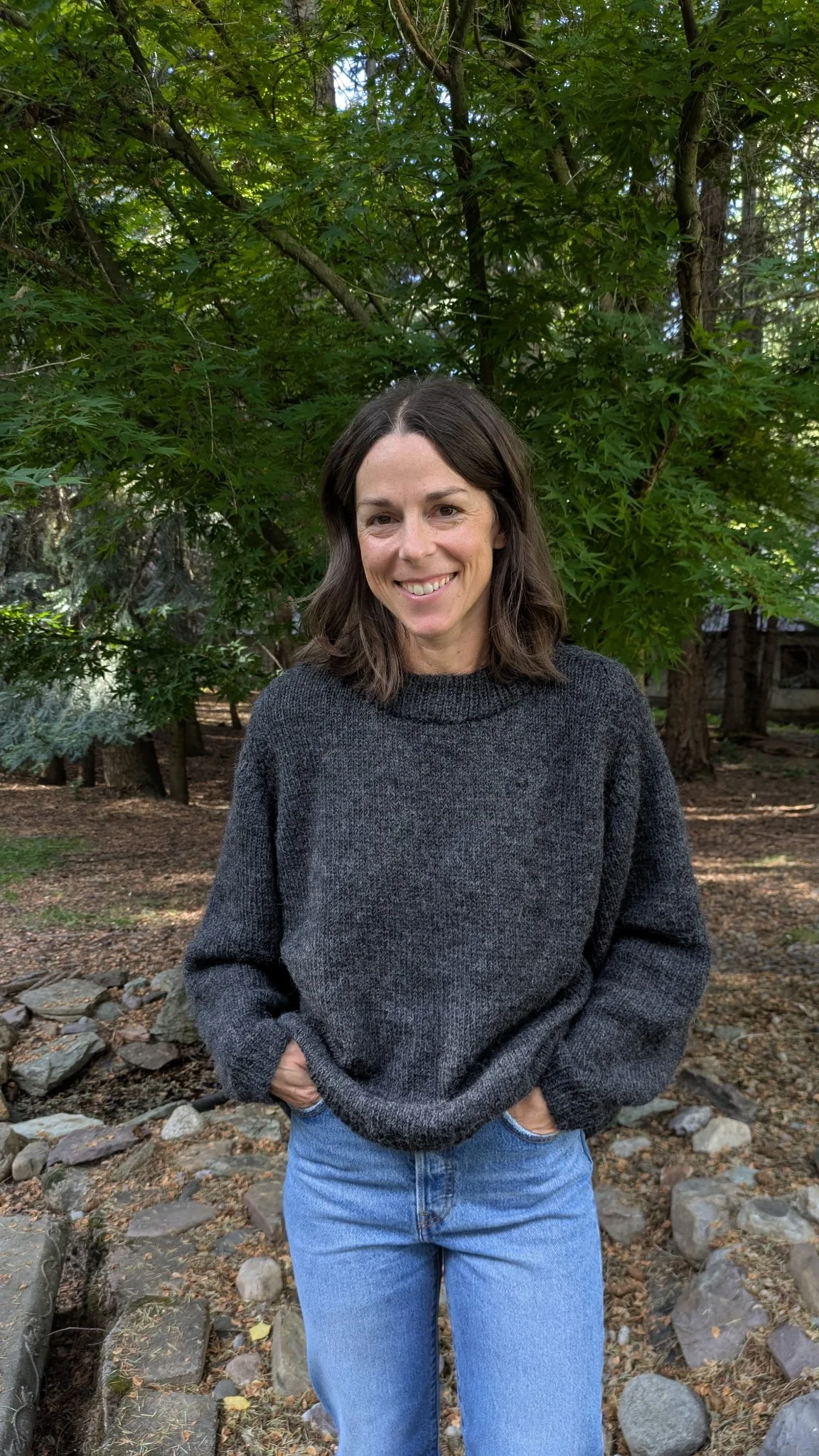 A woman with brown hair, wearing a black sweater and blue jeans, stands outdoors with a smile in front of green trees and rocks.