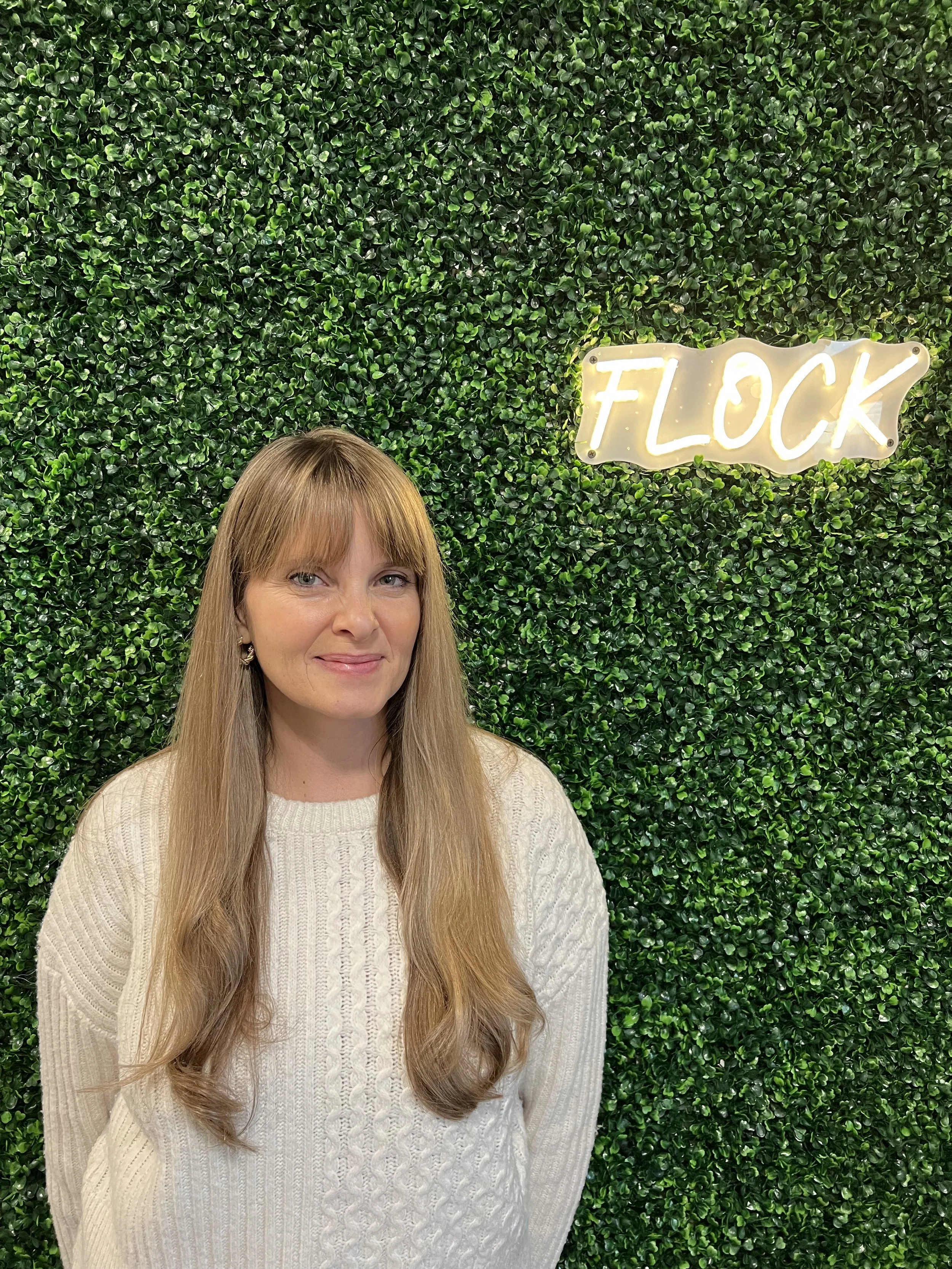 A woman with long, light brown hair and bangs smiling in front of a green artificial hedge wall with a neon sign that says 'FLOCK'.
