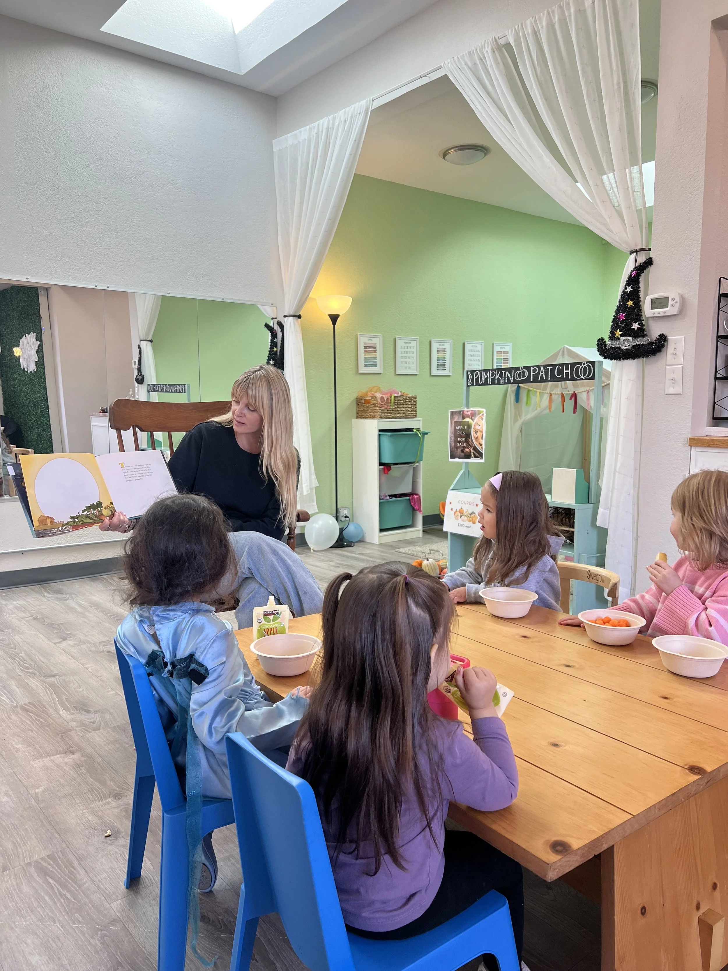 A woman reading a book to five children sitting at a wooden table in a decorated room.