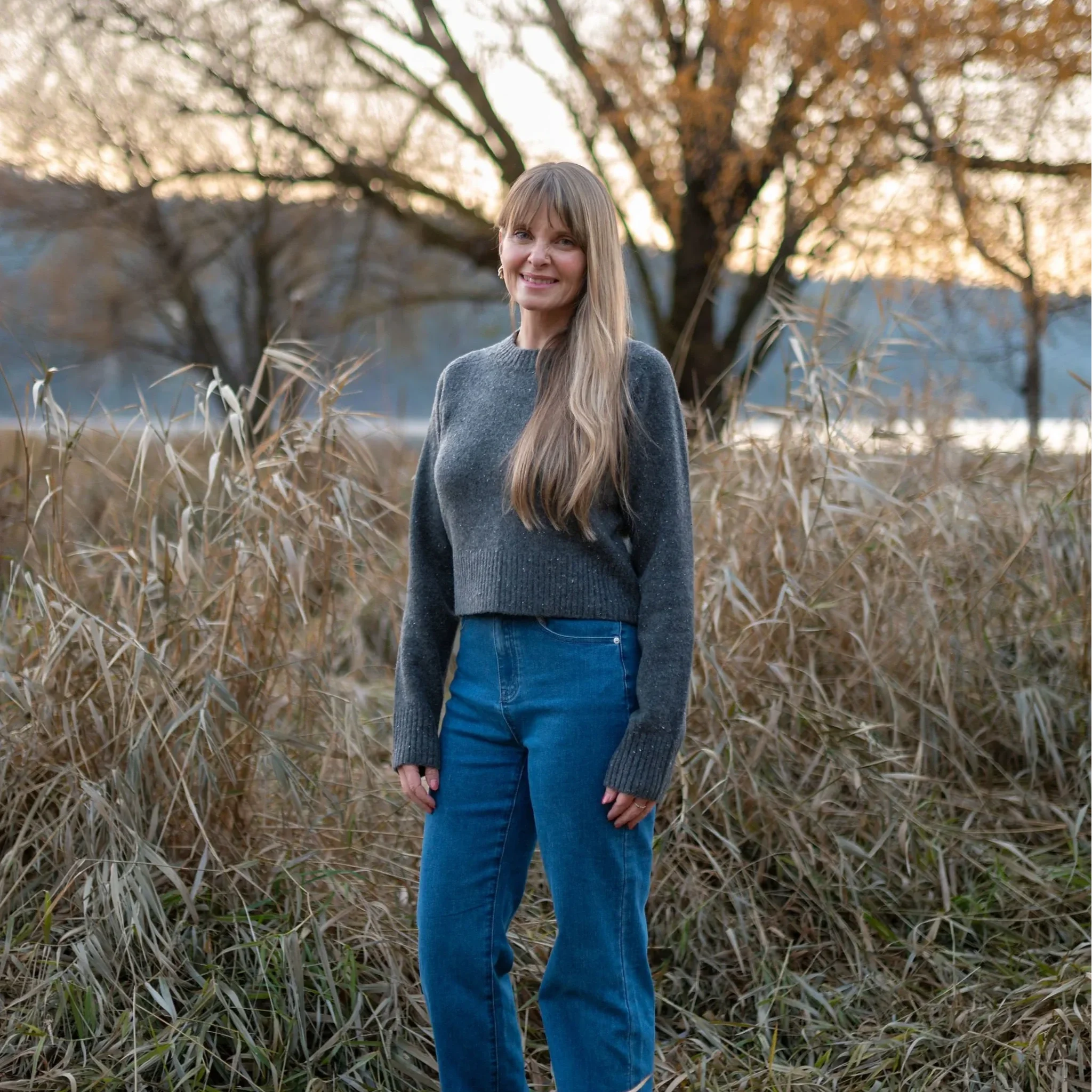 A woman with long blonde hair smiling outdoors during autumn, wearing a gray sweater and blue jeans, standing in front of tall dry grass and a leafless tree.