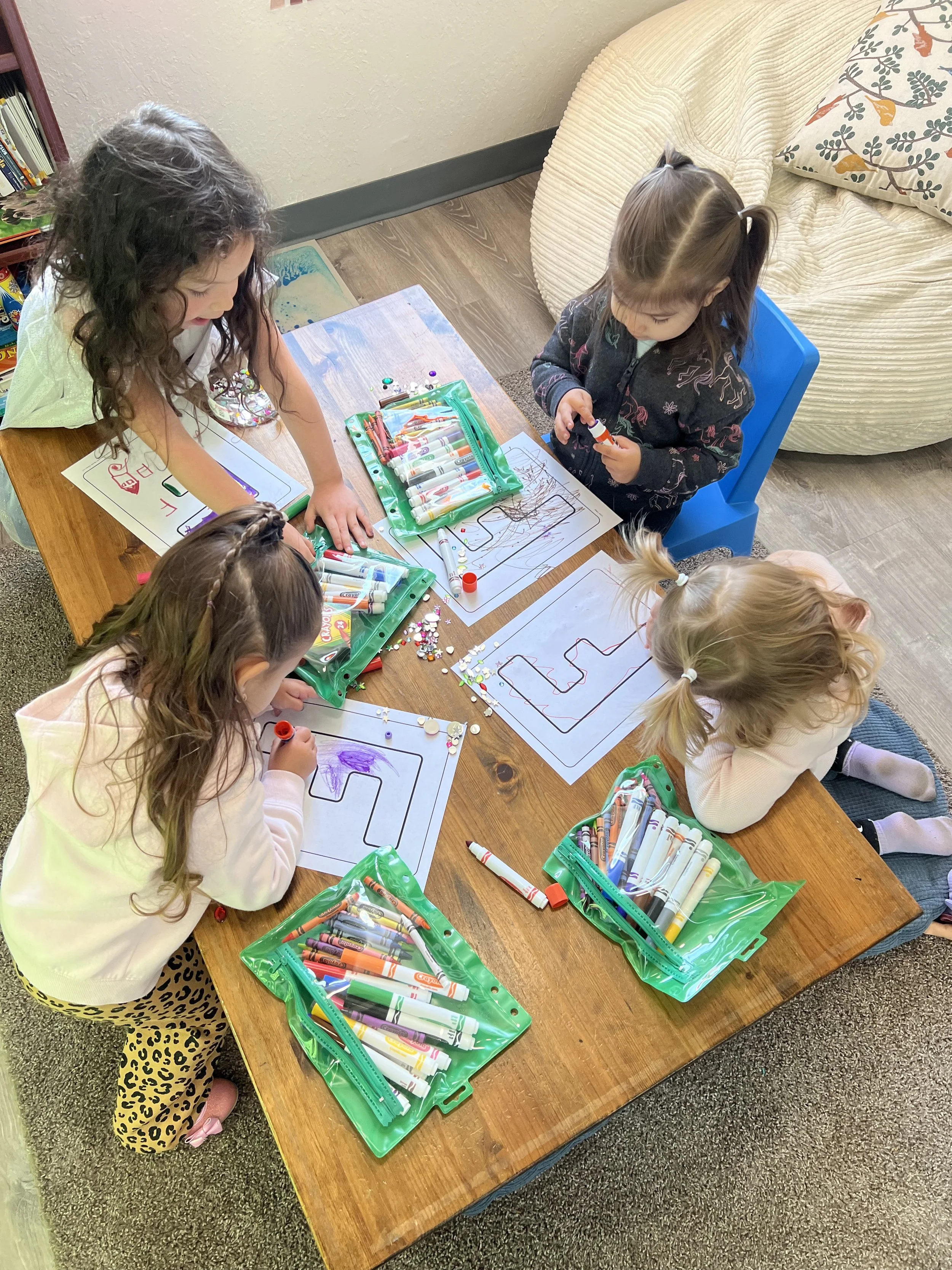 Four young girls are sitting at a wooden table, coloring on sheets of paper with markers and crayons. There are bags of markers on the table, and the girls are focused on their artwork in a cozy room with a sofa and bookshelf.