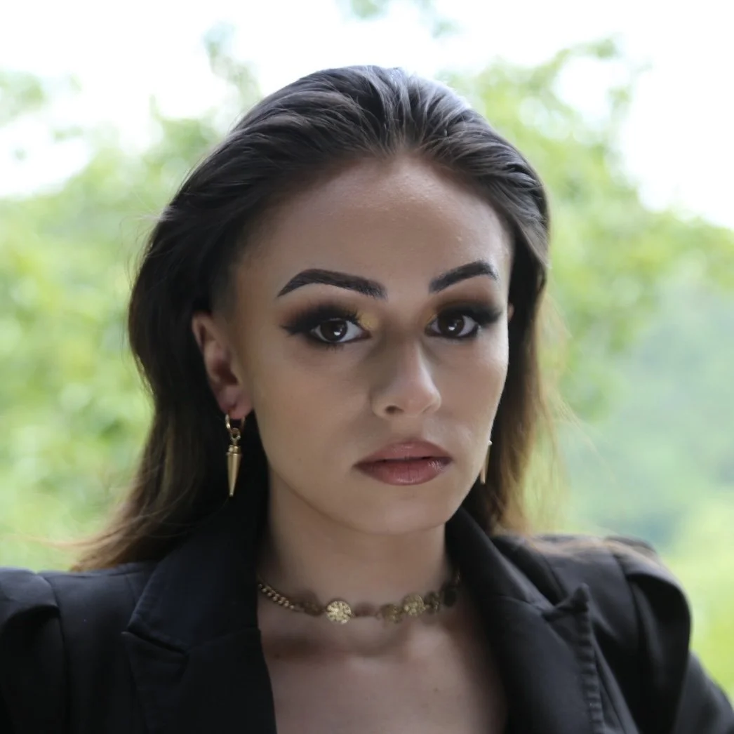 A woman with dark hair, arched eyebrows, and makeup, wearing earrings, a choker necklace, and a black jacket, looks directly at the camera against a blurred green outdoor background.