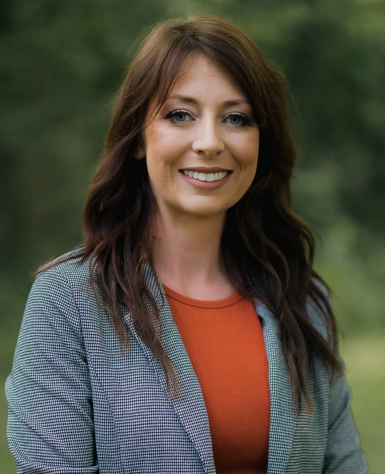 A woman with long brown hair and blue eyes, smiling, outdoors with a blurred green background, wearing a checkered blazer and orange shirt.