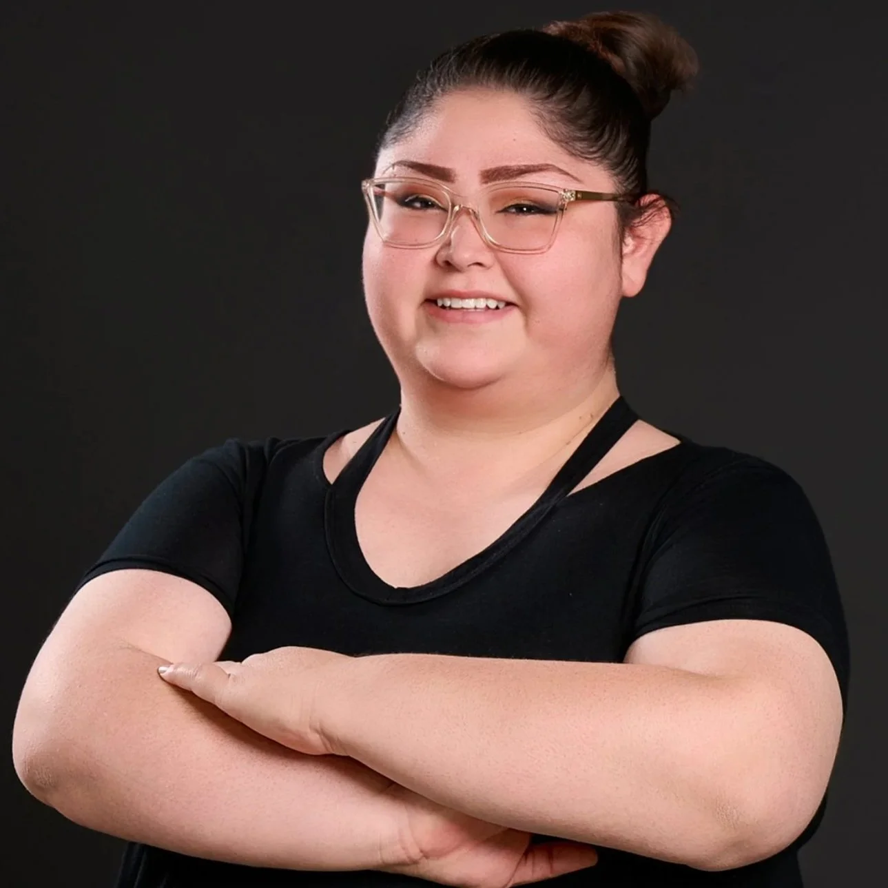 A person with glasses and a top knot hairstyle, smiling with arms crossed, wearing a black t-shirt.